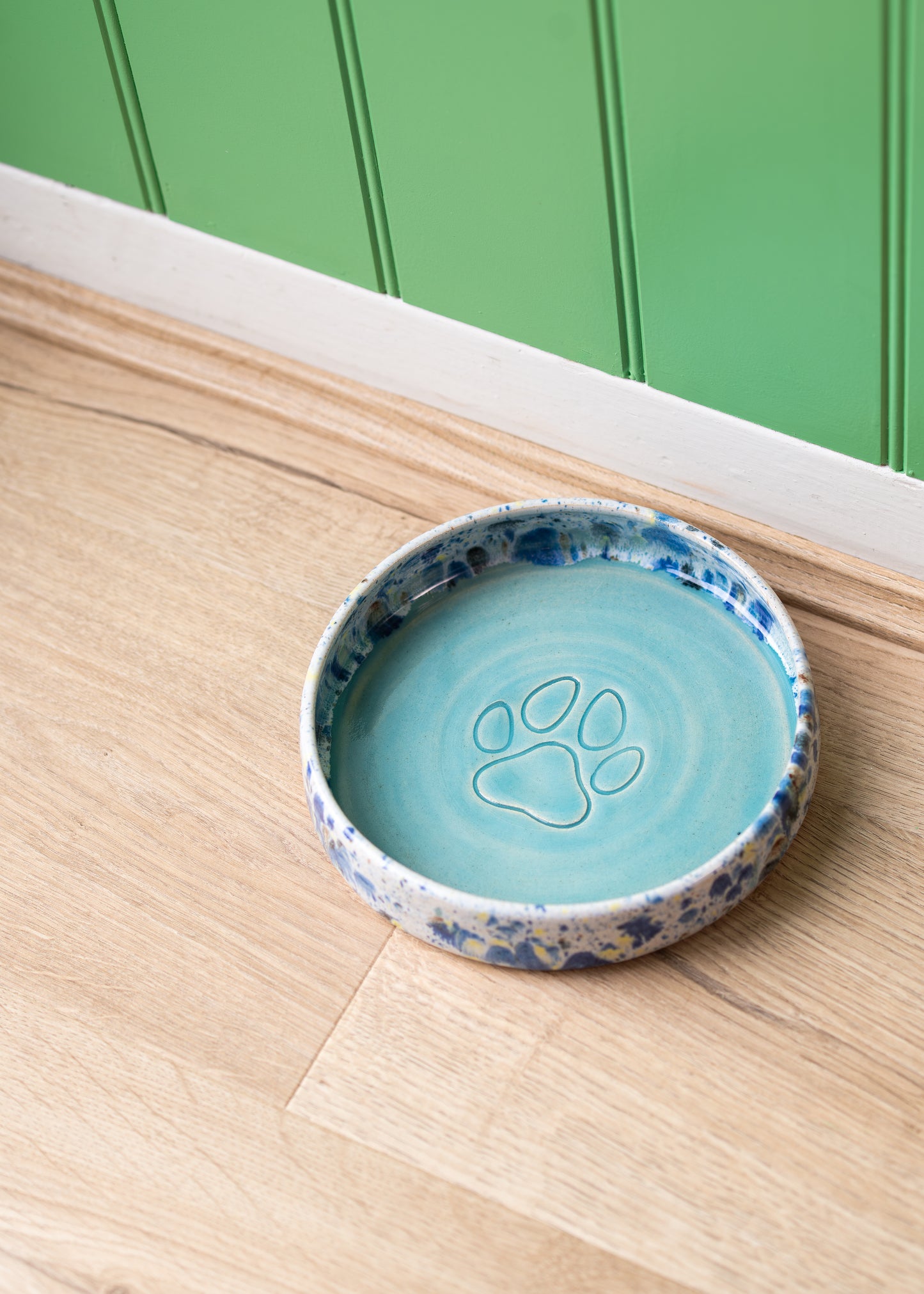 Ceramic pet bowl with blue interior and splatter exterior on a wooden surface with green paneling in the background.
