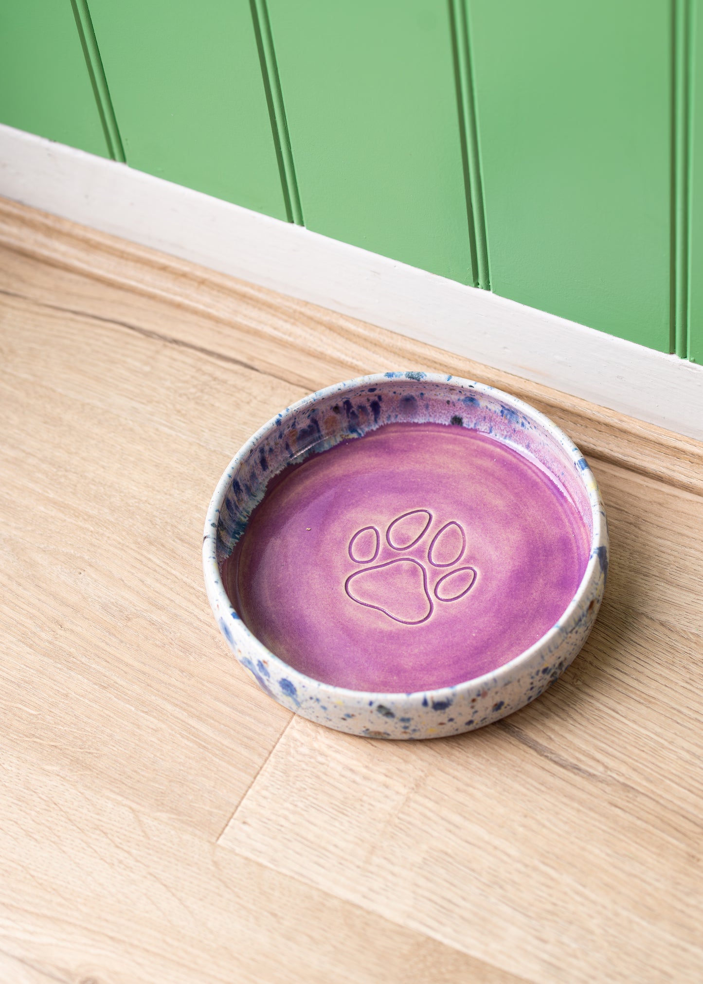 Purple ceramic pet bowl with paw print design on a wooden surface against a green wall.