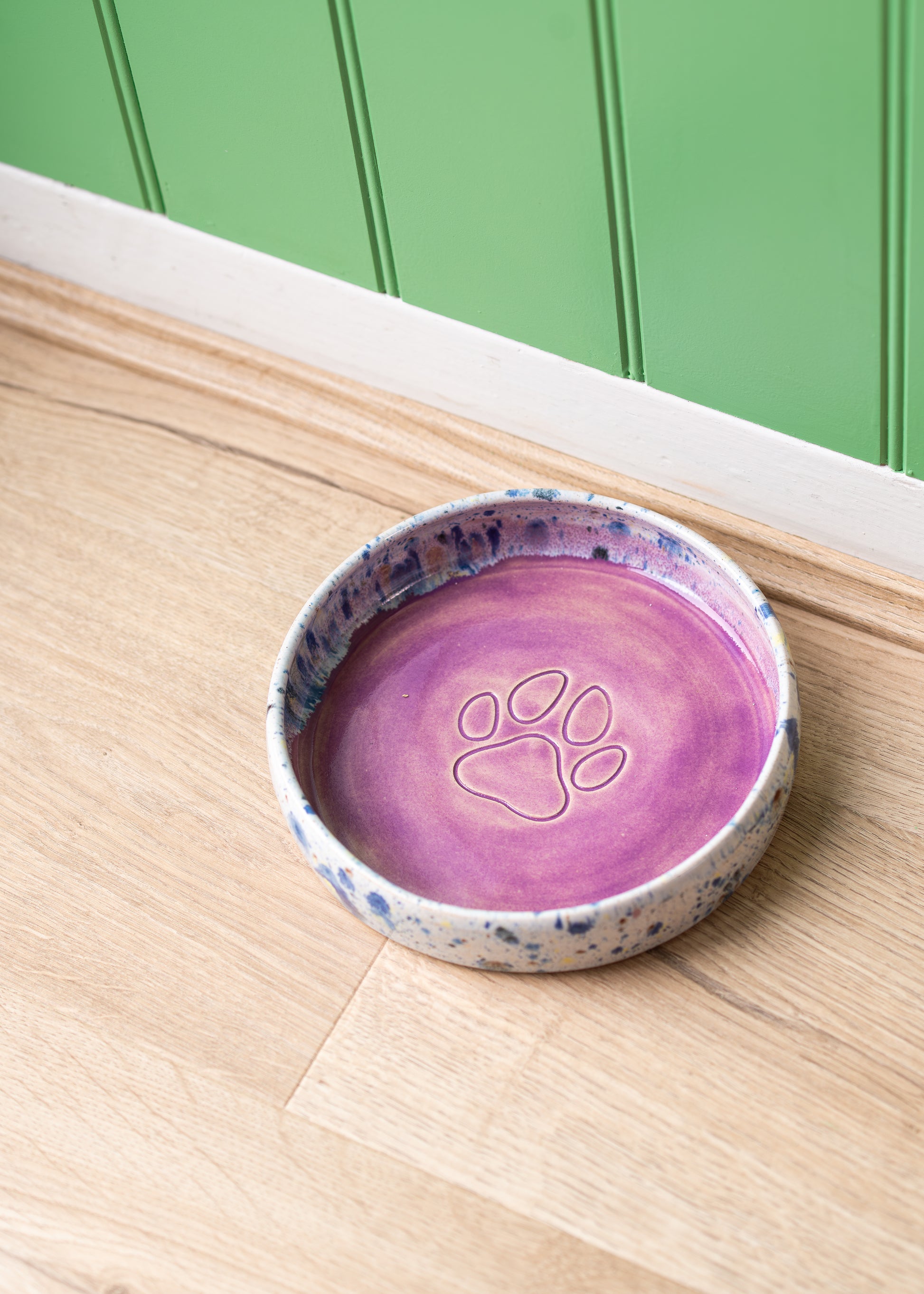 Purple ceramic pet bowl with paw print design on a wooden surface against a green wall.