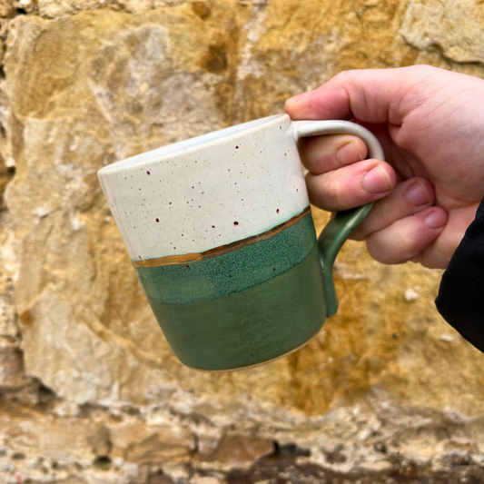 Hand holding a ceramic mug with a speckled top and green bottom against a stone wall background