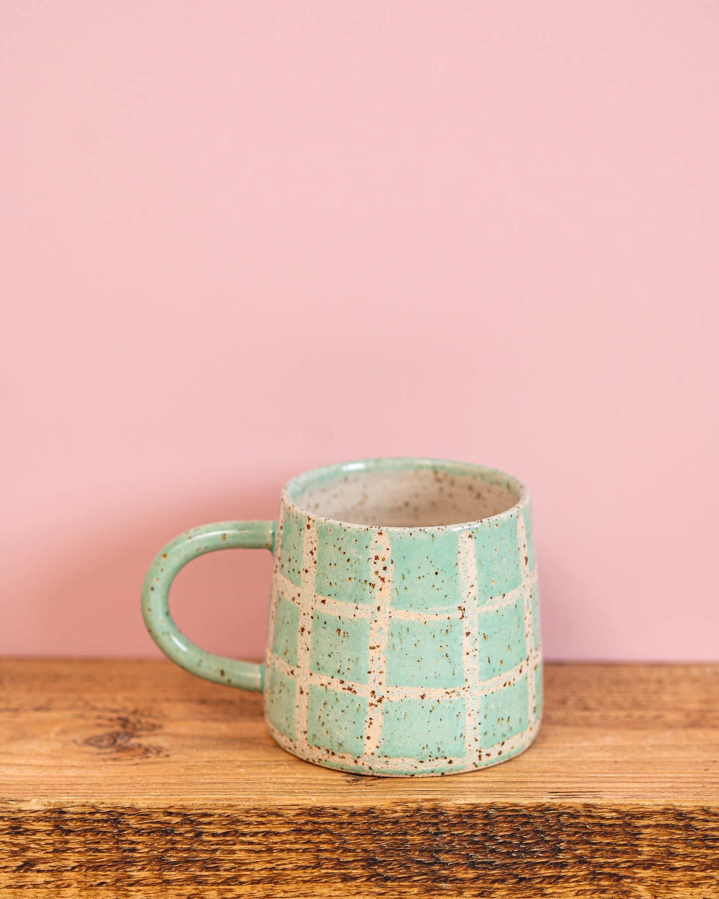 Green ceramic mug with checkered pattern on a wooden surface and pink background