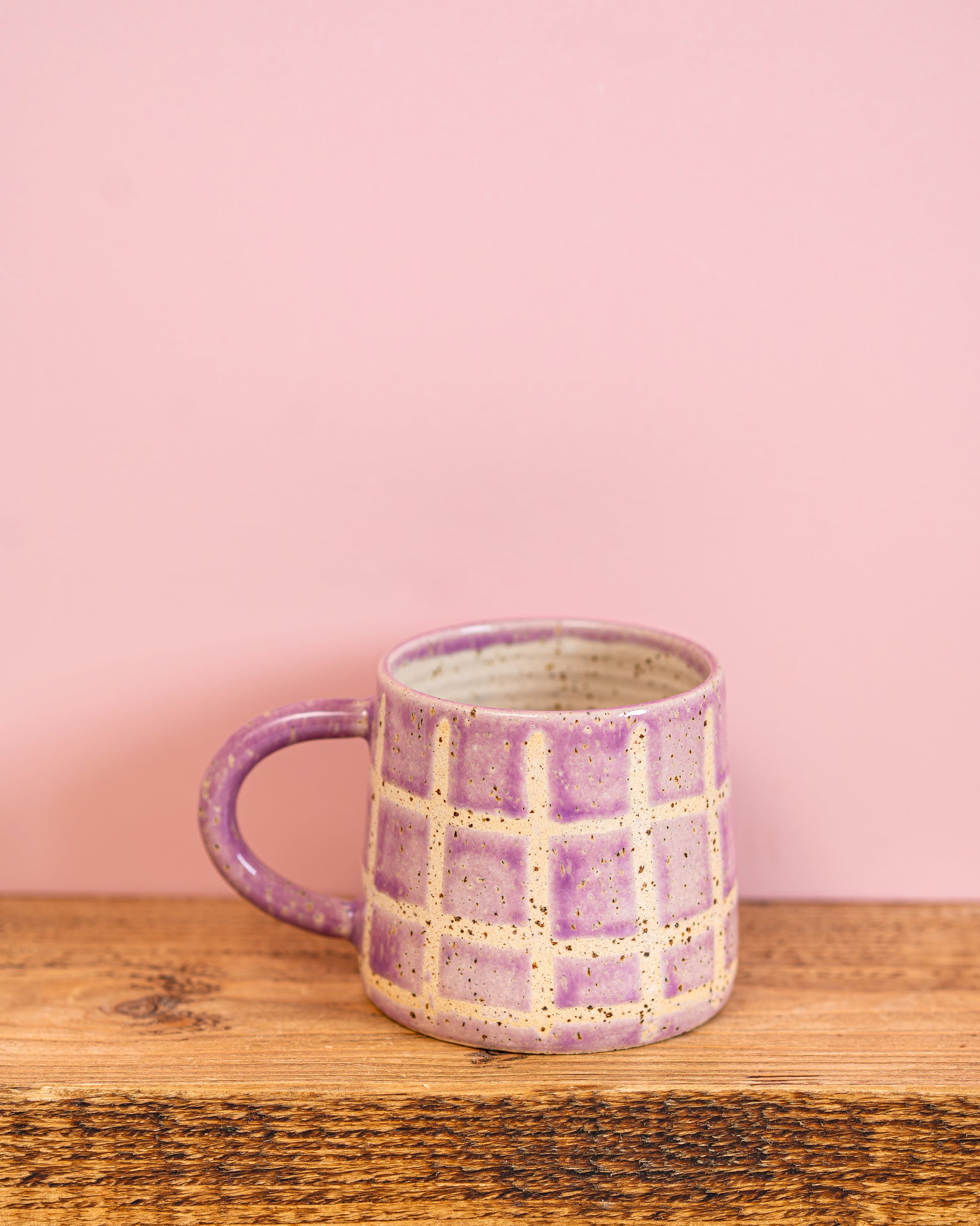 Purple ceramic mug with checkered pattern on a wooden surface and pink background