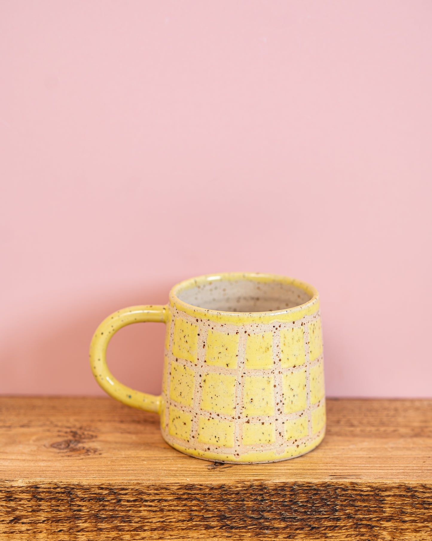 Yellow ceramic mug with speckled pattern on a wooden surface against a pink background
