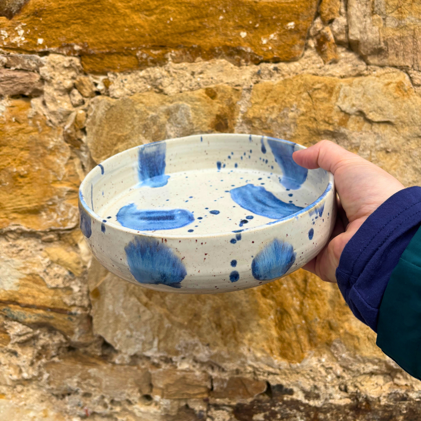 Hand holding a ceramic bowl with blue speckles against a stone wall background
