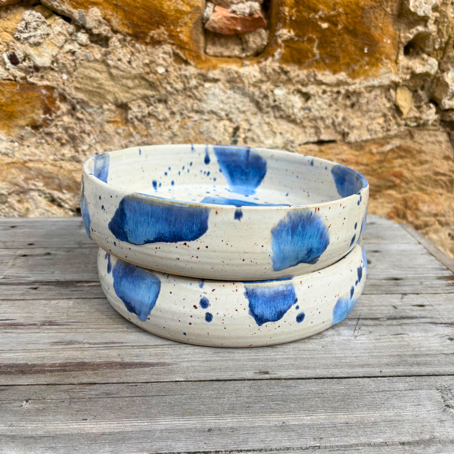 Two ceramic bowls with blue speckled design on a wooden surface with a stone wall background.