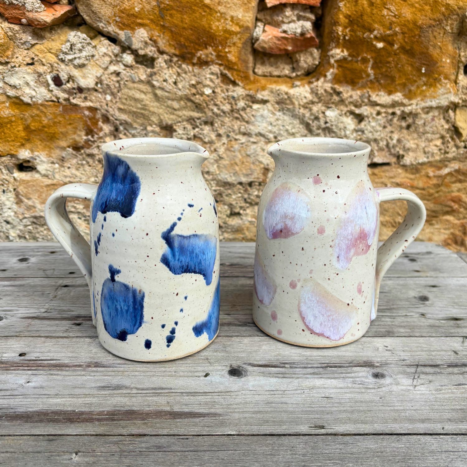 Two ceramic pitchers with abstract patterns on a wooden surface against a stone wall.