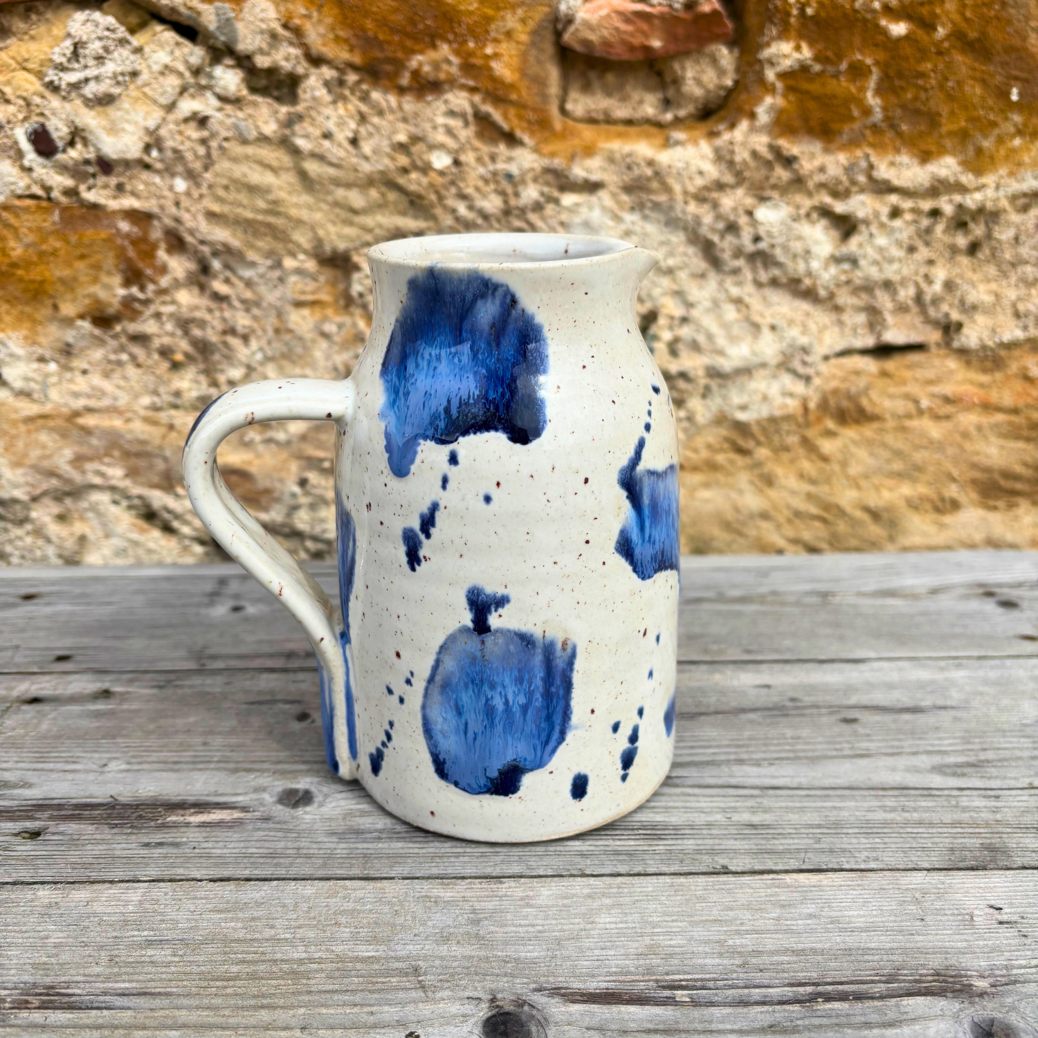 Ceramic pitcher with blue splashes on a wooden surface with a stone wall background
