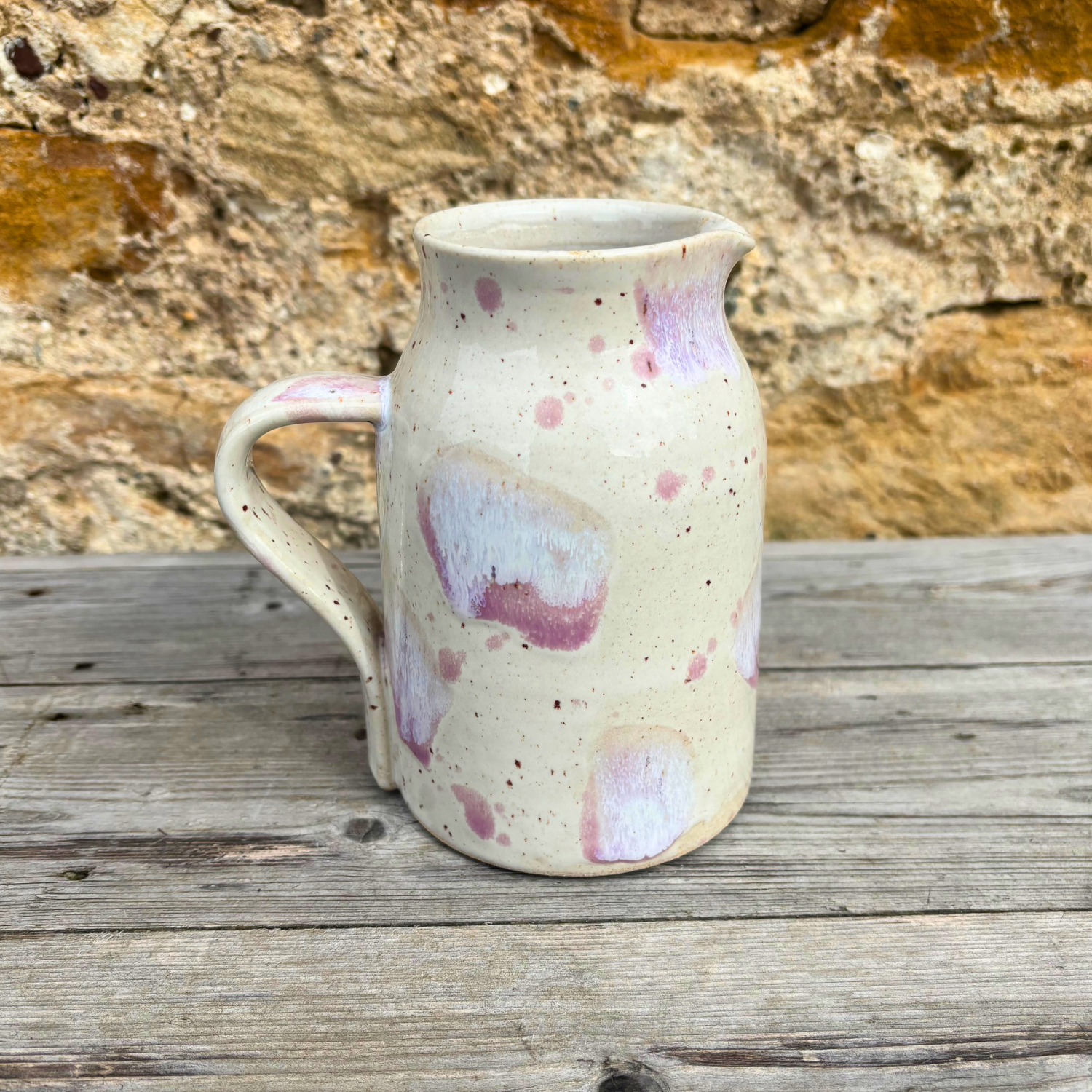 Speckled ceramic pitcher on a wooden surface with a stone wall background