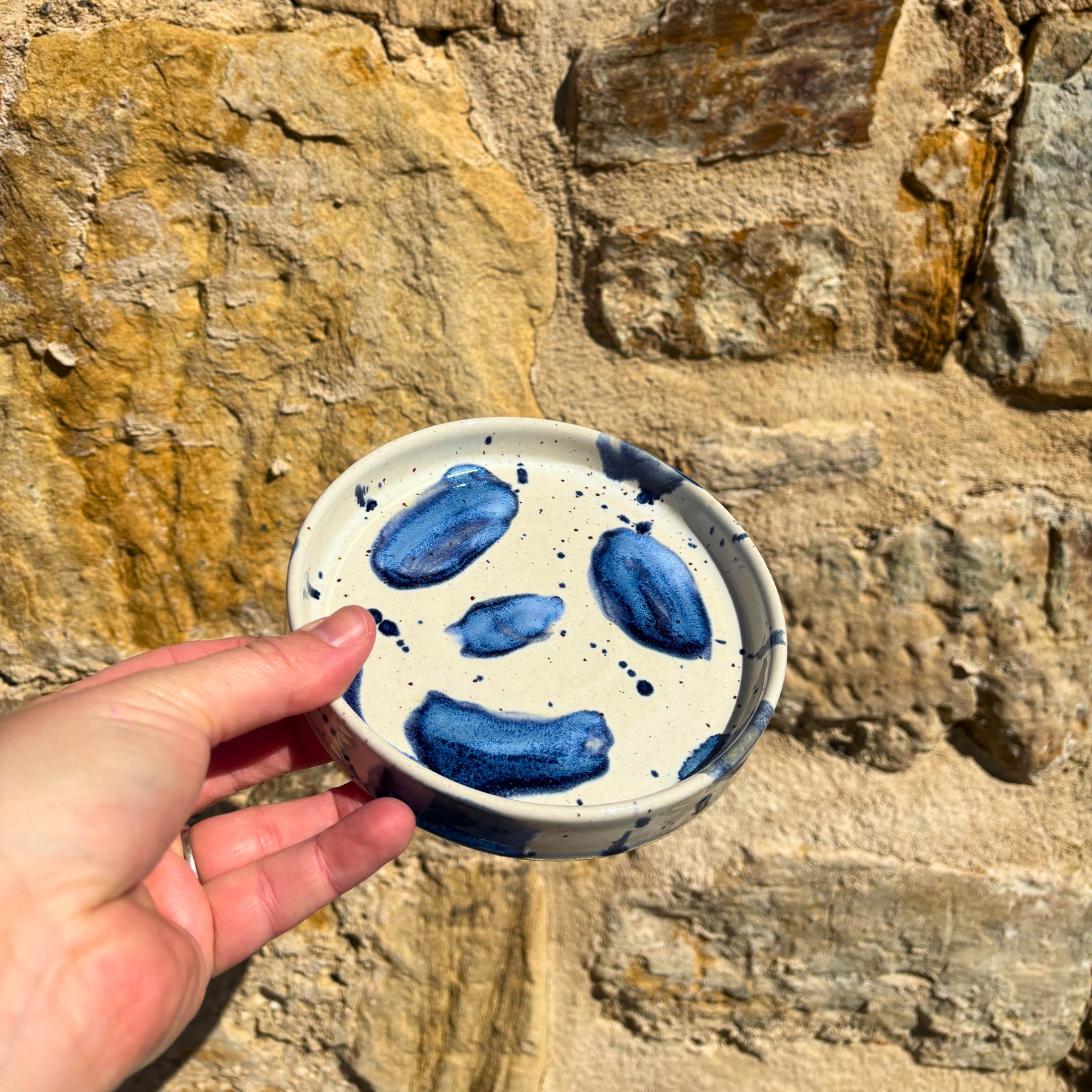 Hand holding a small ceramic trinket dish with blue speckles against a stone wall background