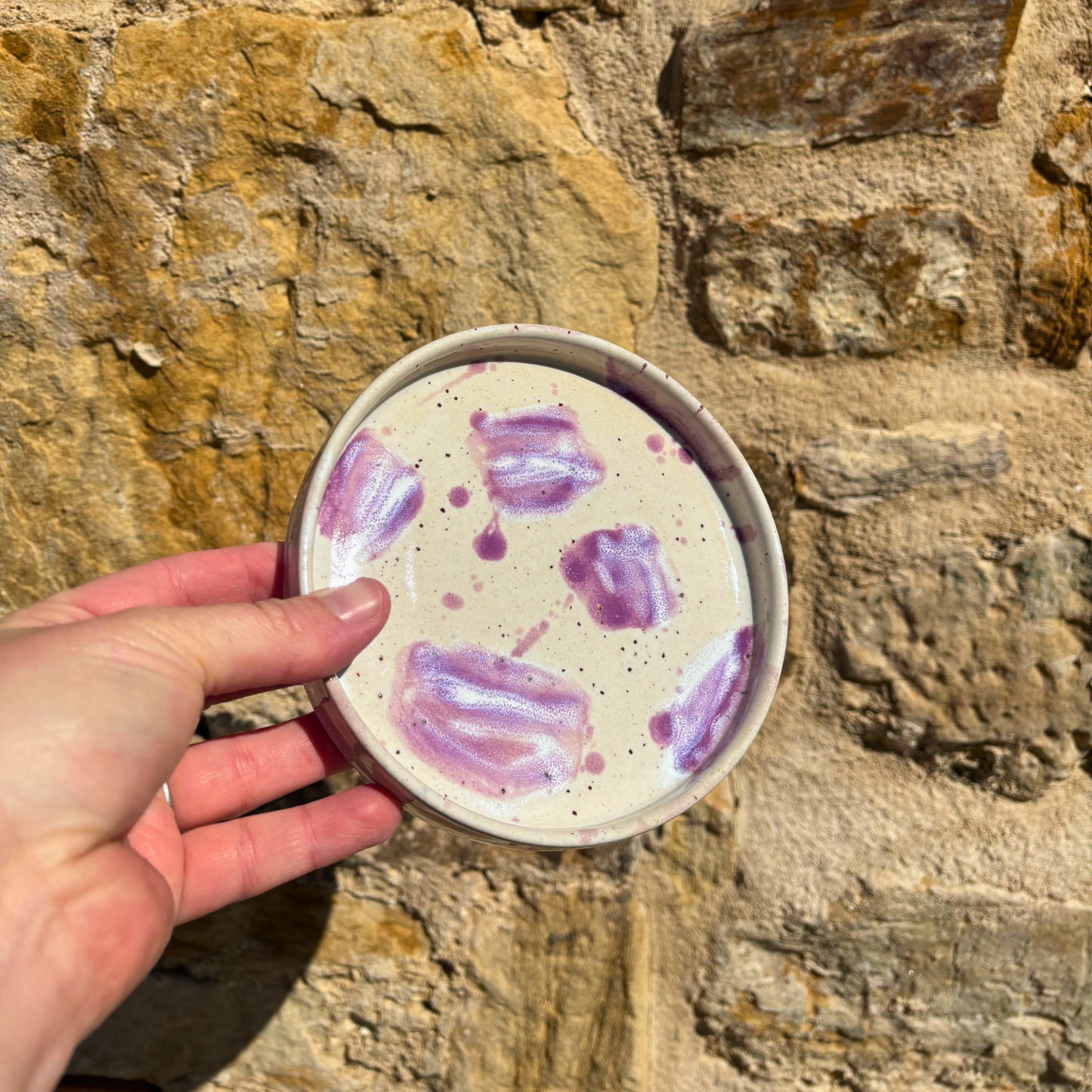 Hand holding a trinket dish with purple designs against a stone wall background