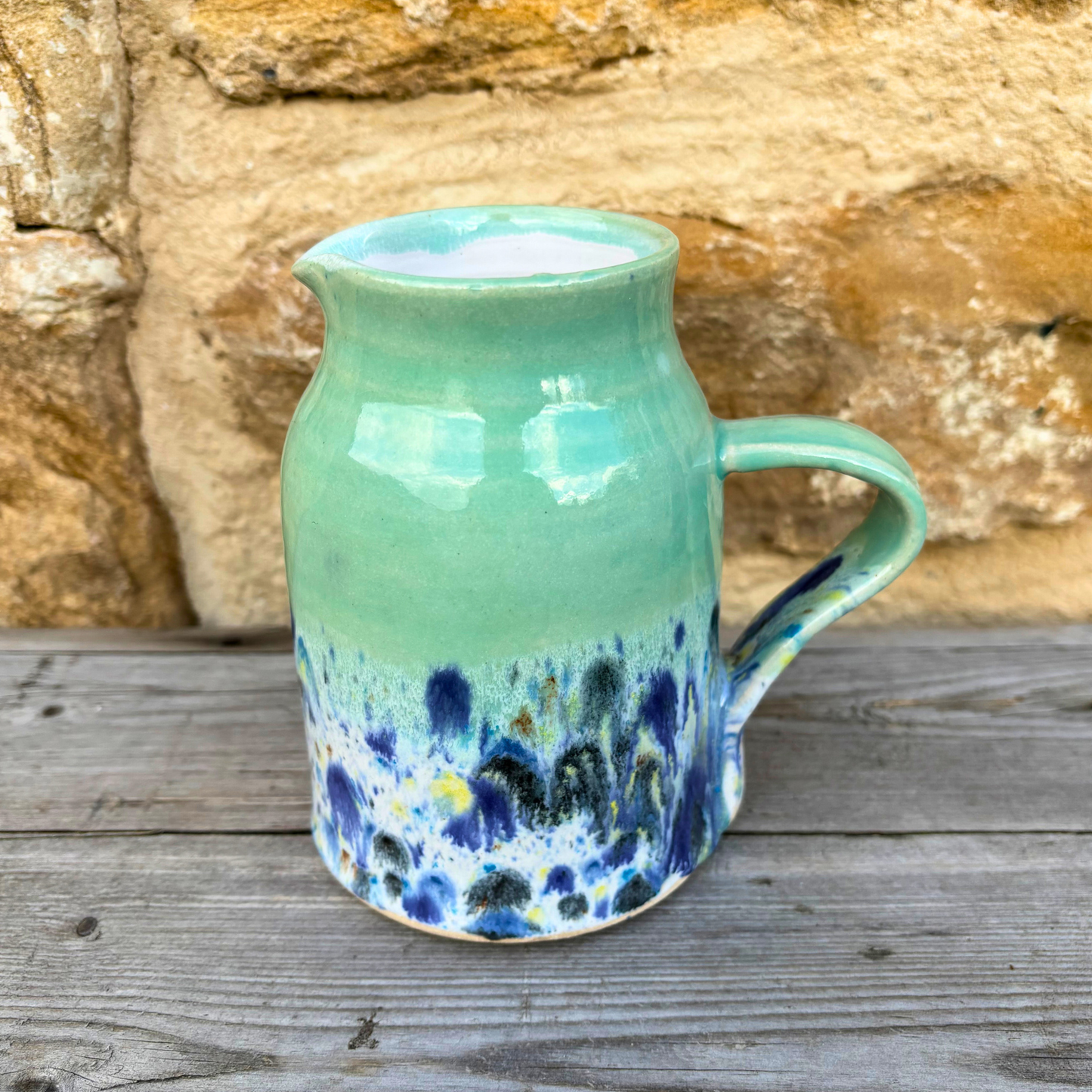 Green ceramic jug with blue and yellow speckles on a wooden surface with stone wall background