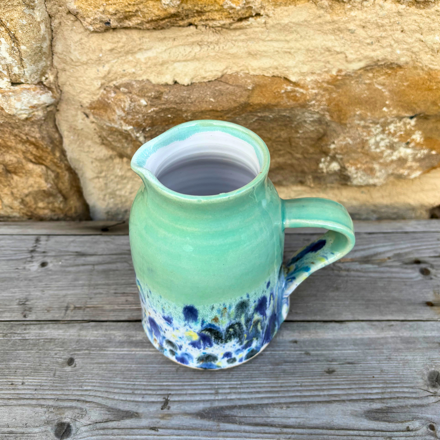 Green ceramic jug with blue and white speckled design on a wooden surface with stone wall background