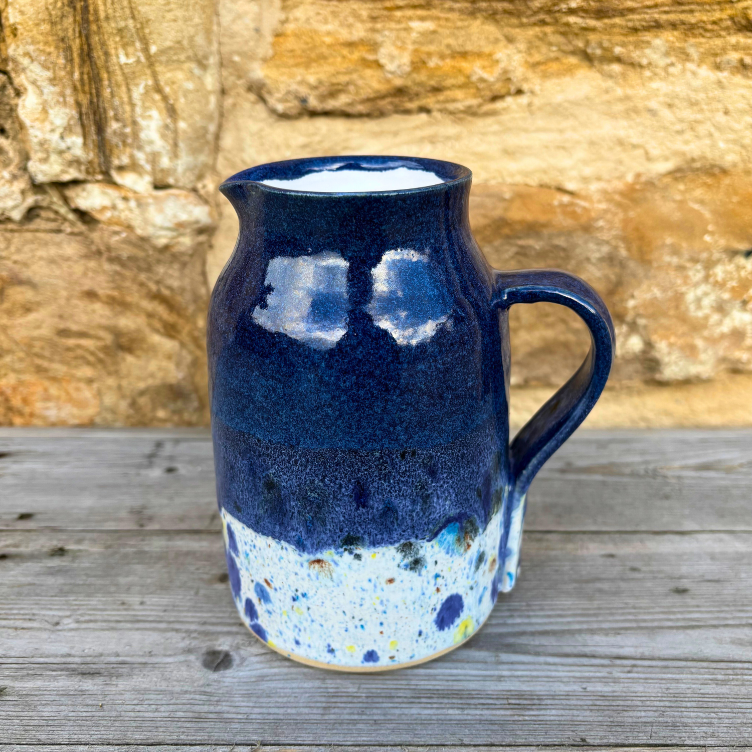 Blue ceramic pitcher with speckled design on a wooden surface with stone wall background