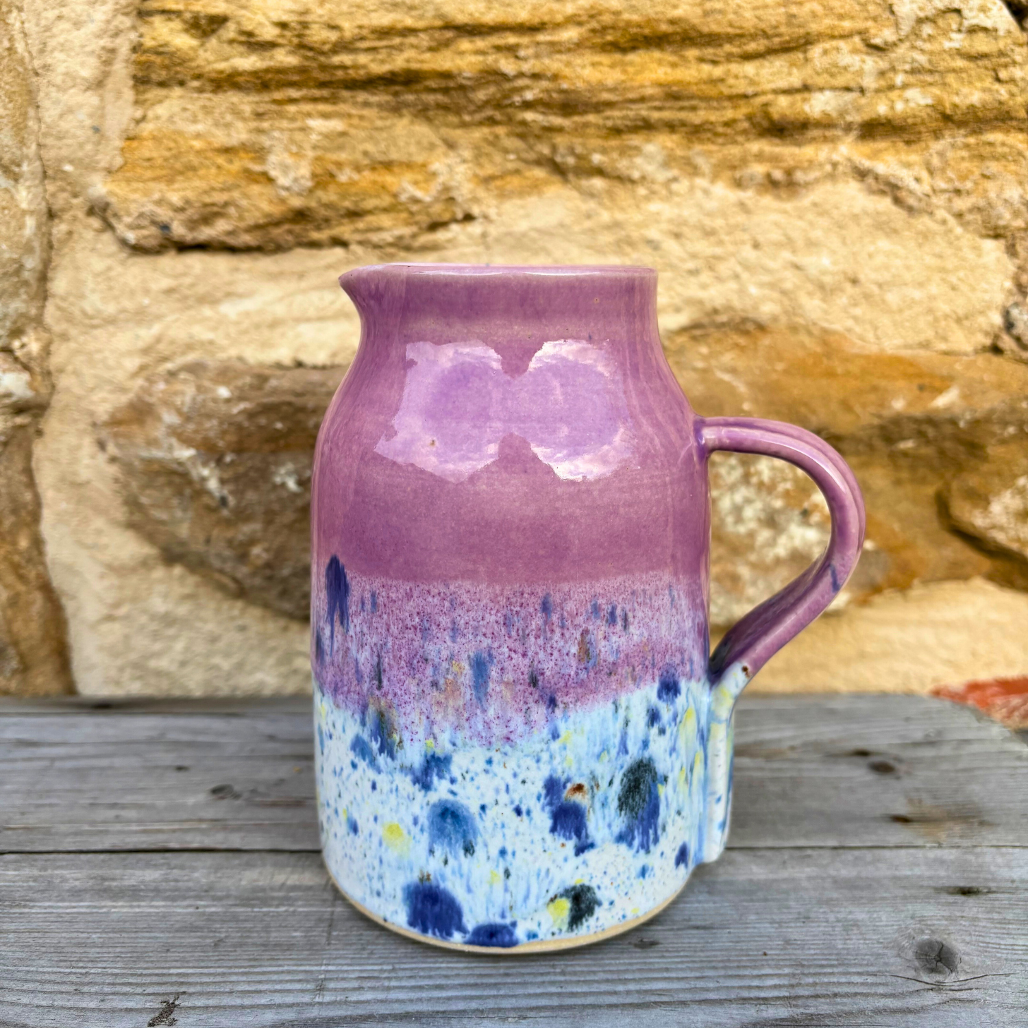 Pink ceramic pitcher with blue speckled design on a wooden surface with stone background