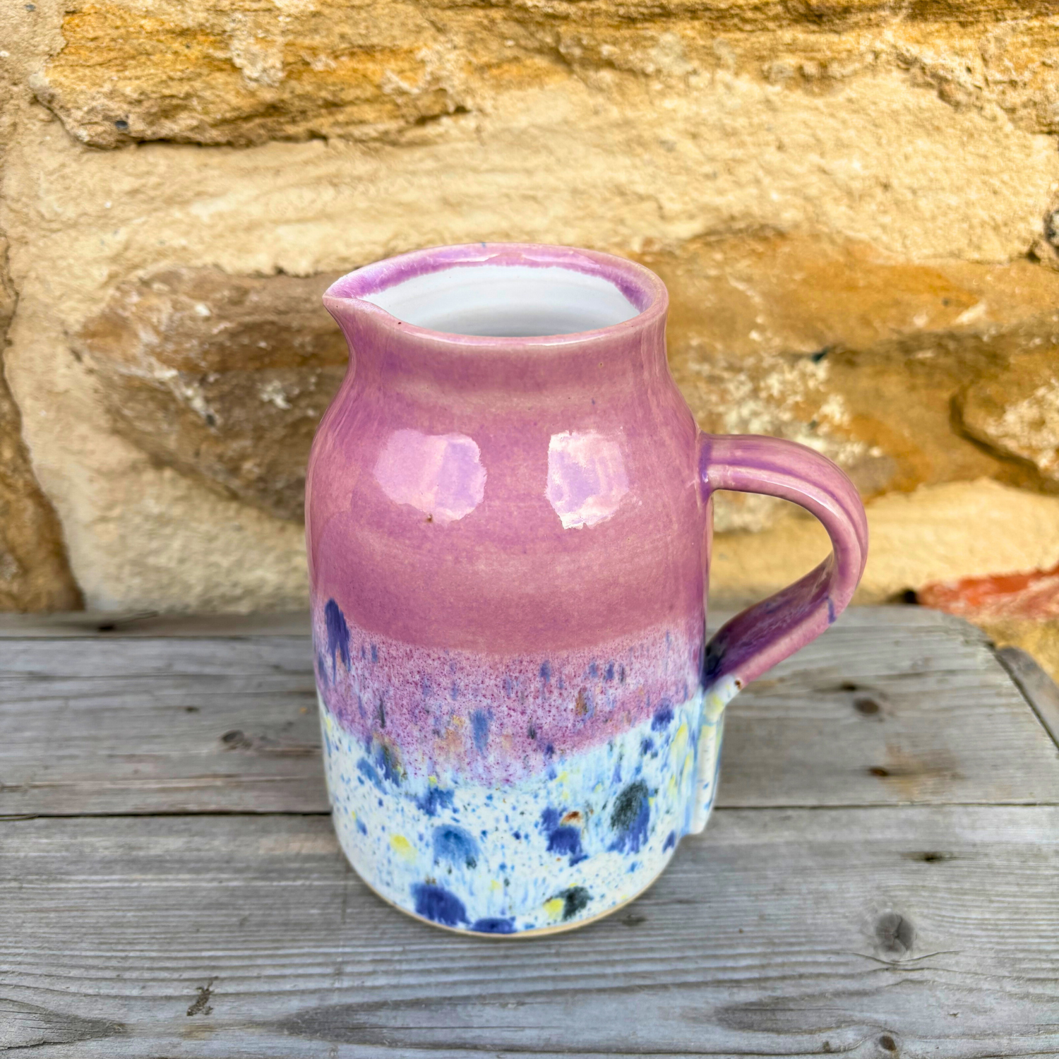 Pink ceramic pitcher with speckled design on a wooden surface with stone wall background