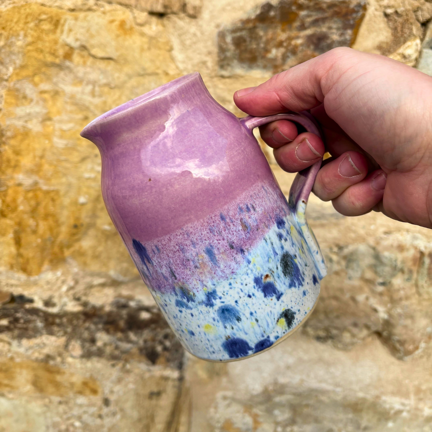 Hand holding a pink and blue speckled ceramic mug against a stone wall background