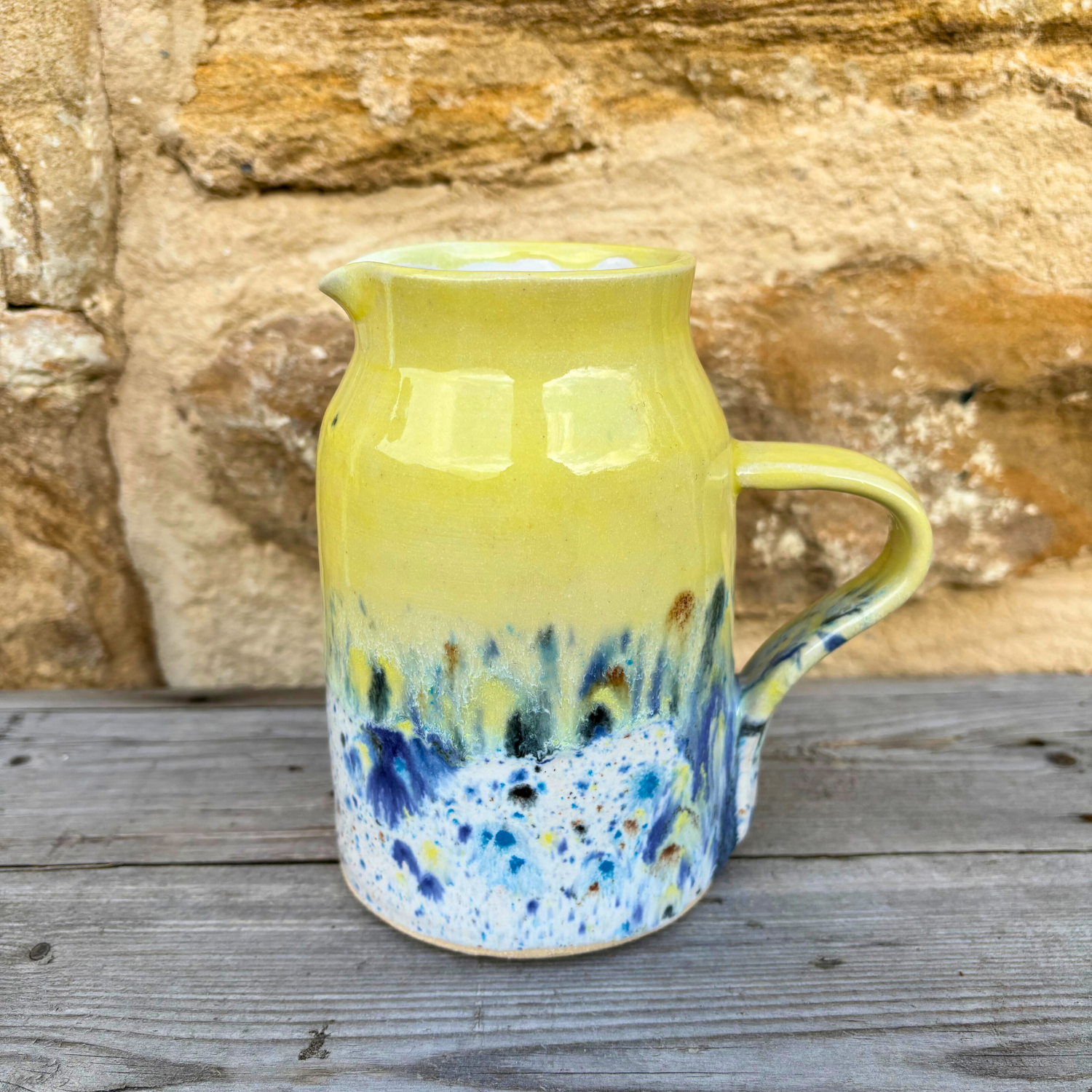 Yellow ceramic jug with blue speckles on a wooden surface with a stone wall background