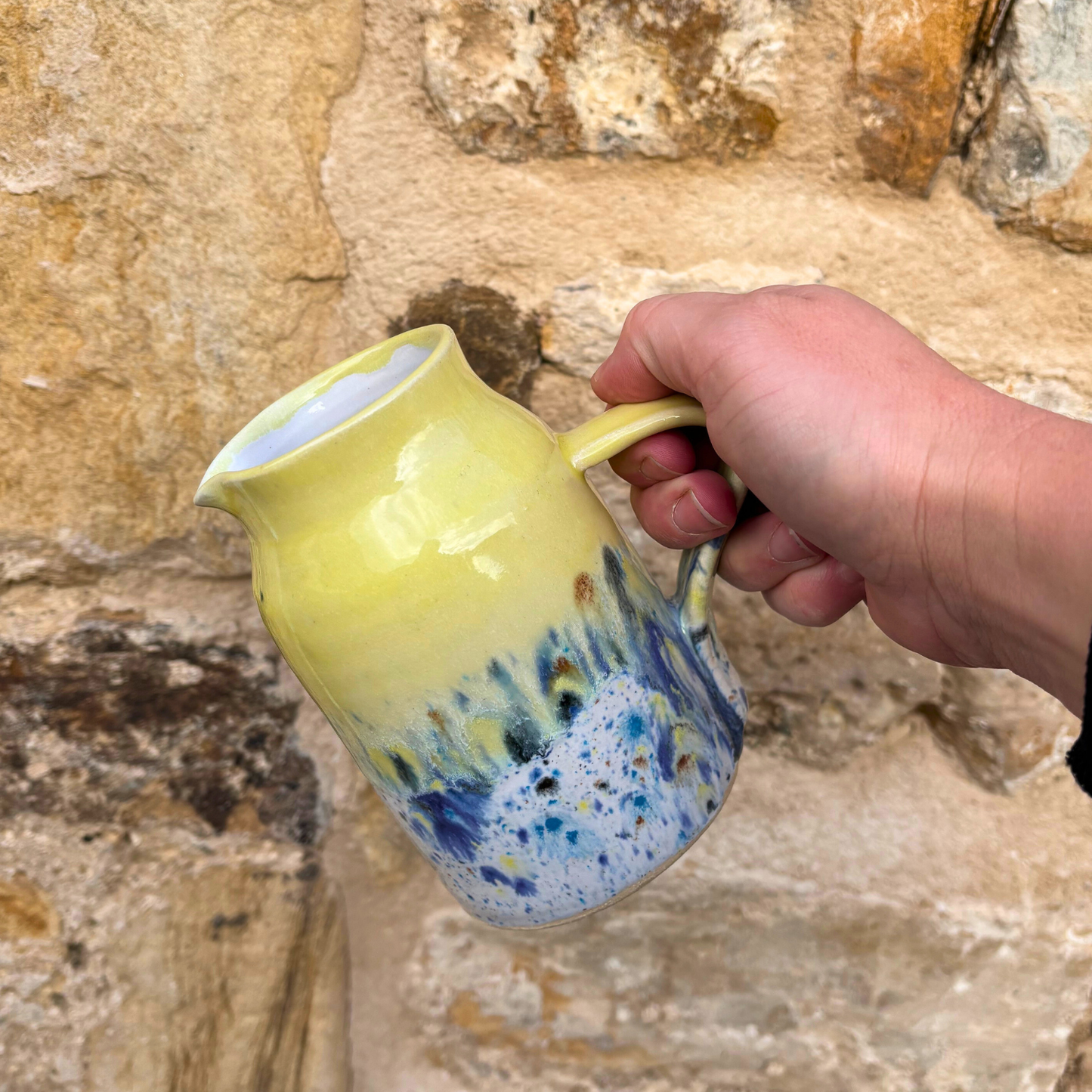 Hand holding a ceramic pitcher with a colorful design against a stone wall.