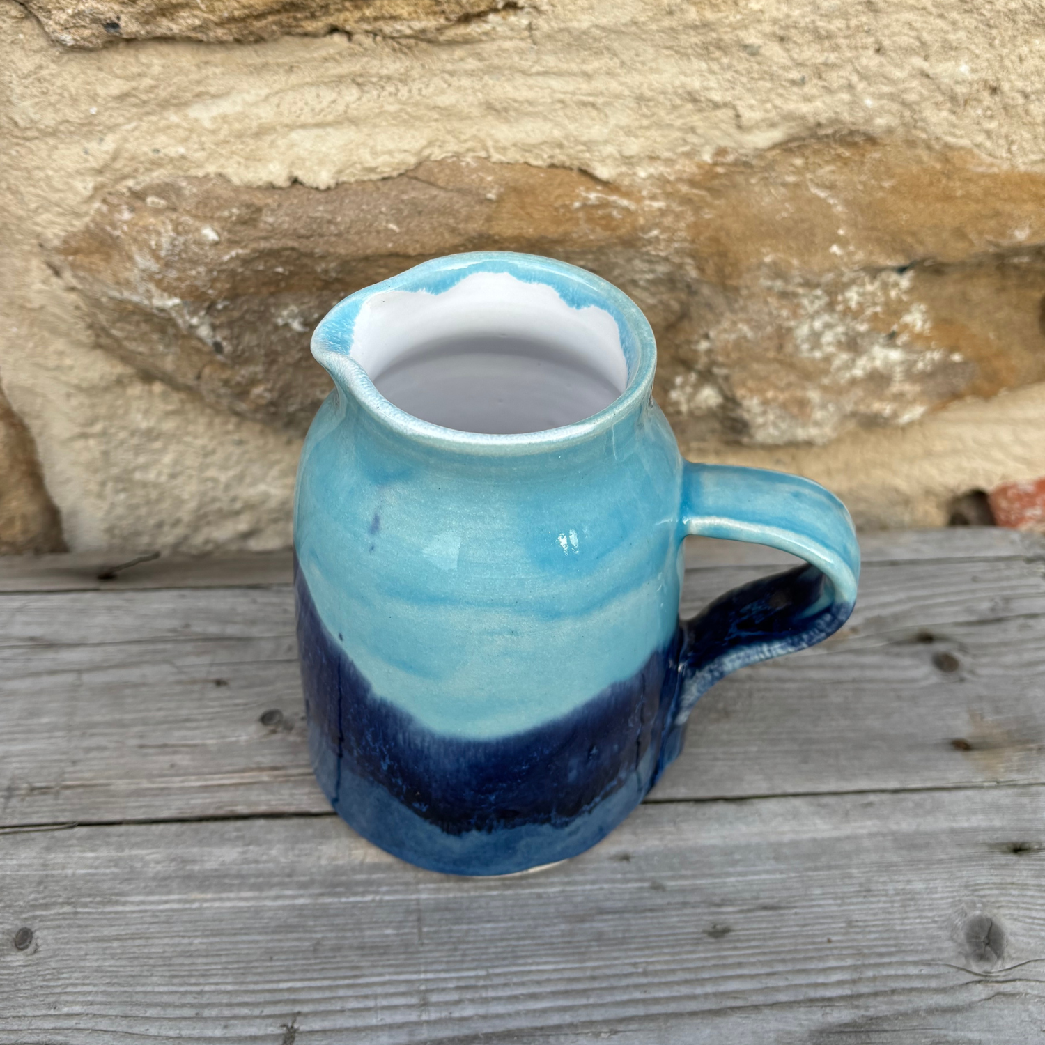 Blue ceramic pitcher with a white interior on a wooden surface with a stone background