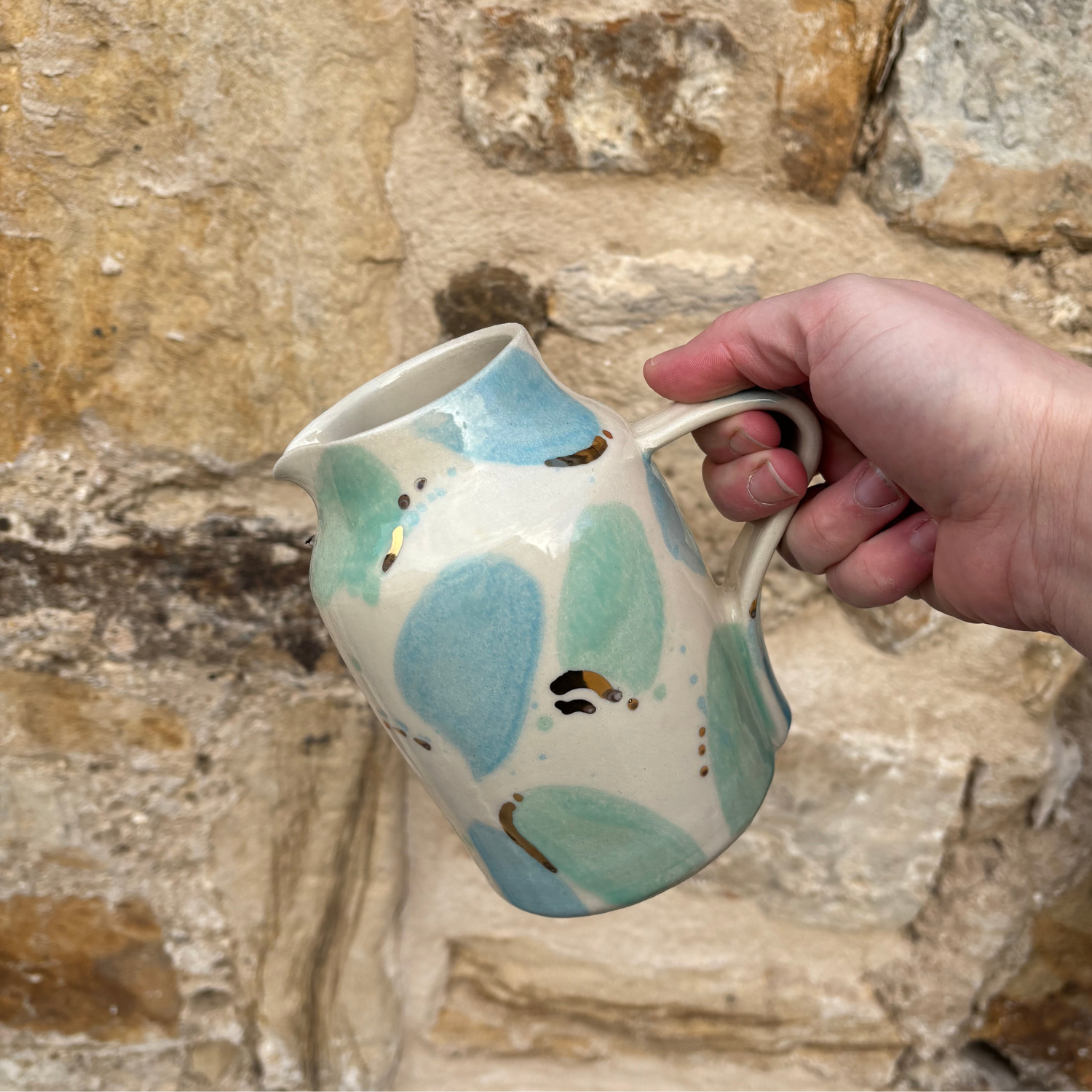 Hand holding a ceramic pitcher with blue and green spots against a stone wall background