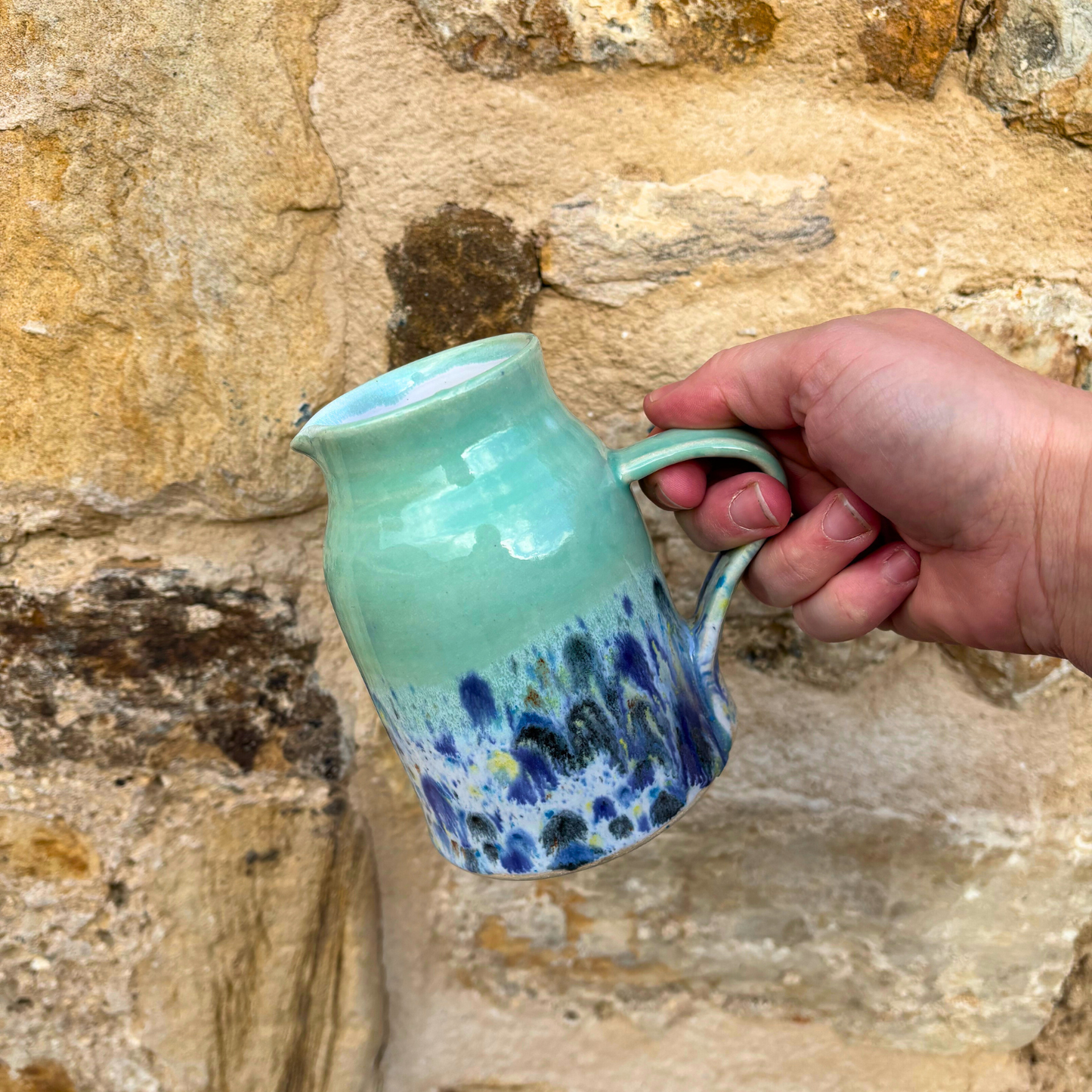 Hand holding a green ceramic jug with blue speckles against a stone wall background
