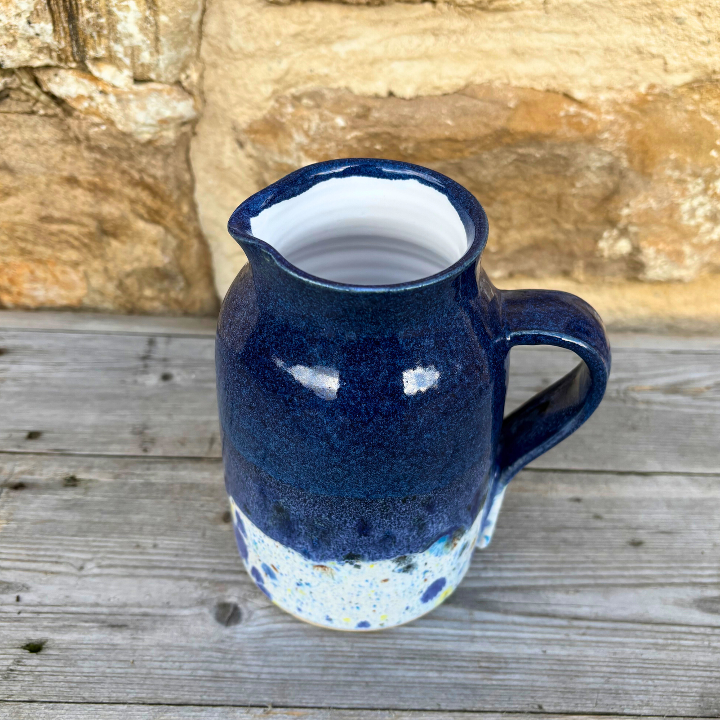 Blue ceramic pitcher with a speckled base on a wooden surface with a stone wall background