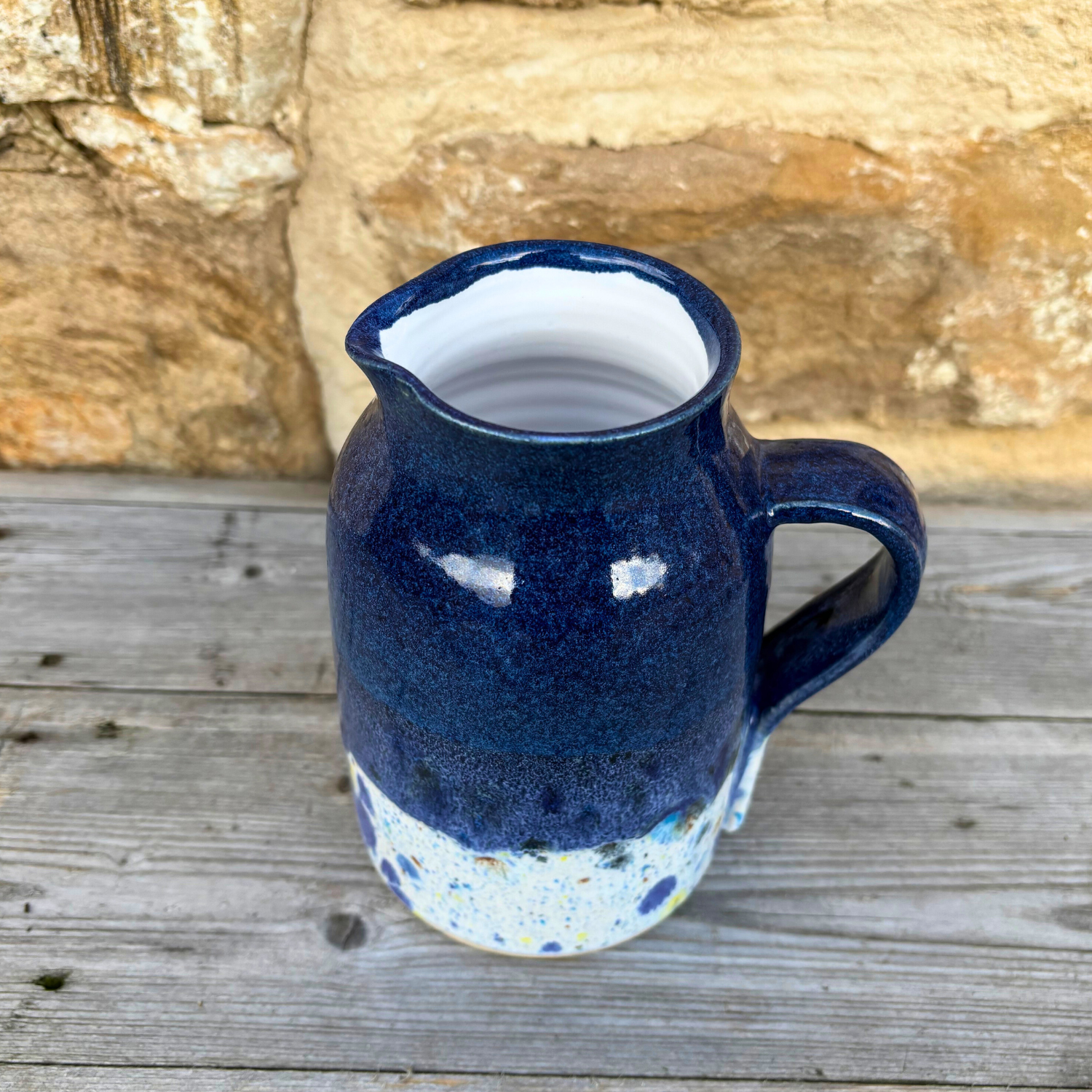 Blue ceramic pitcher with a speckled base on a wooden surface with a stone wall background