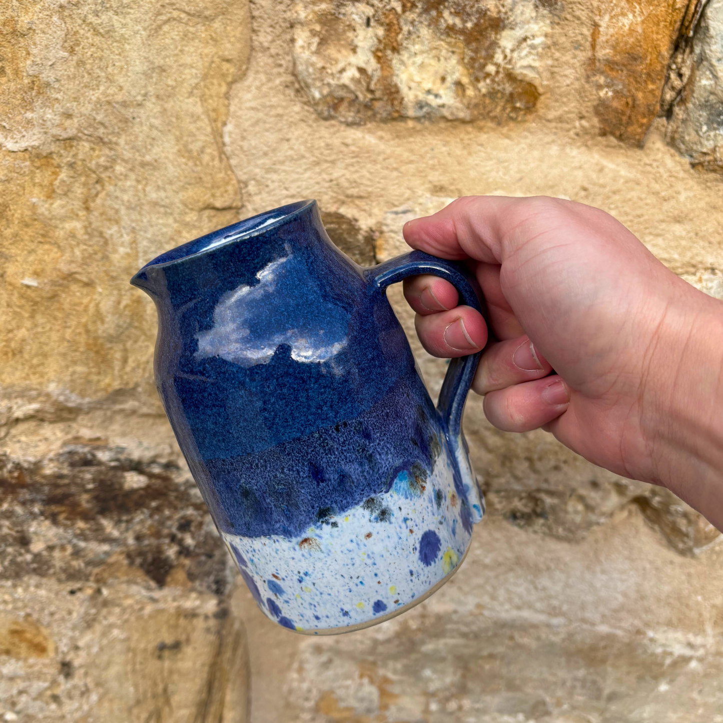 Hand holding a blue ceramic jug against a stone wall background