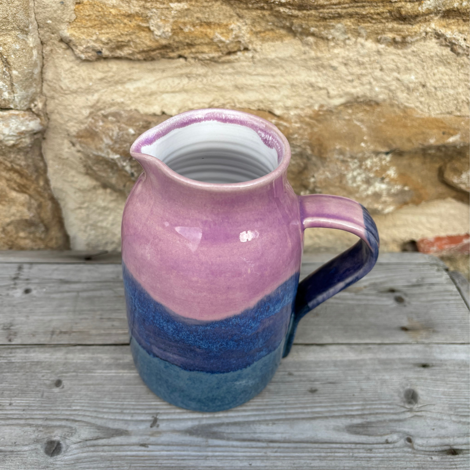 Ceramic pitcher with pink and blue gradient on a wooden surface with stone wall background