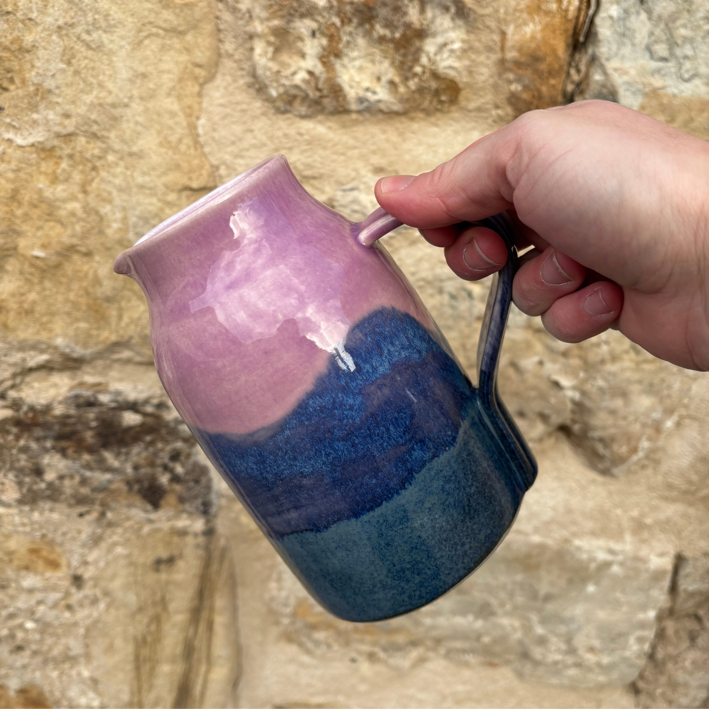Hand holding a ceramic mug with pink and blue gradient against a stone wall background