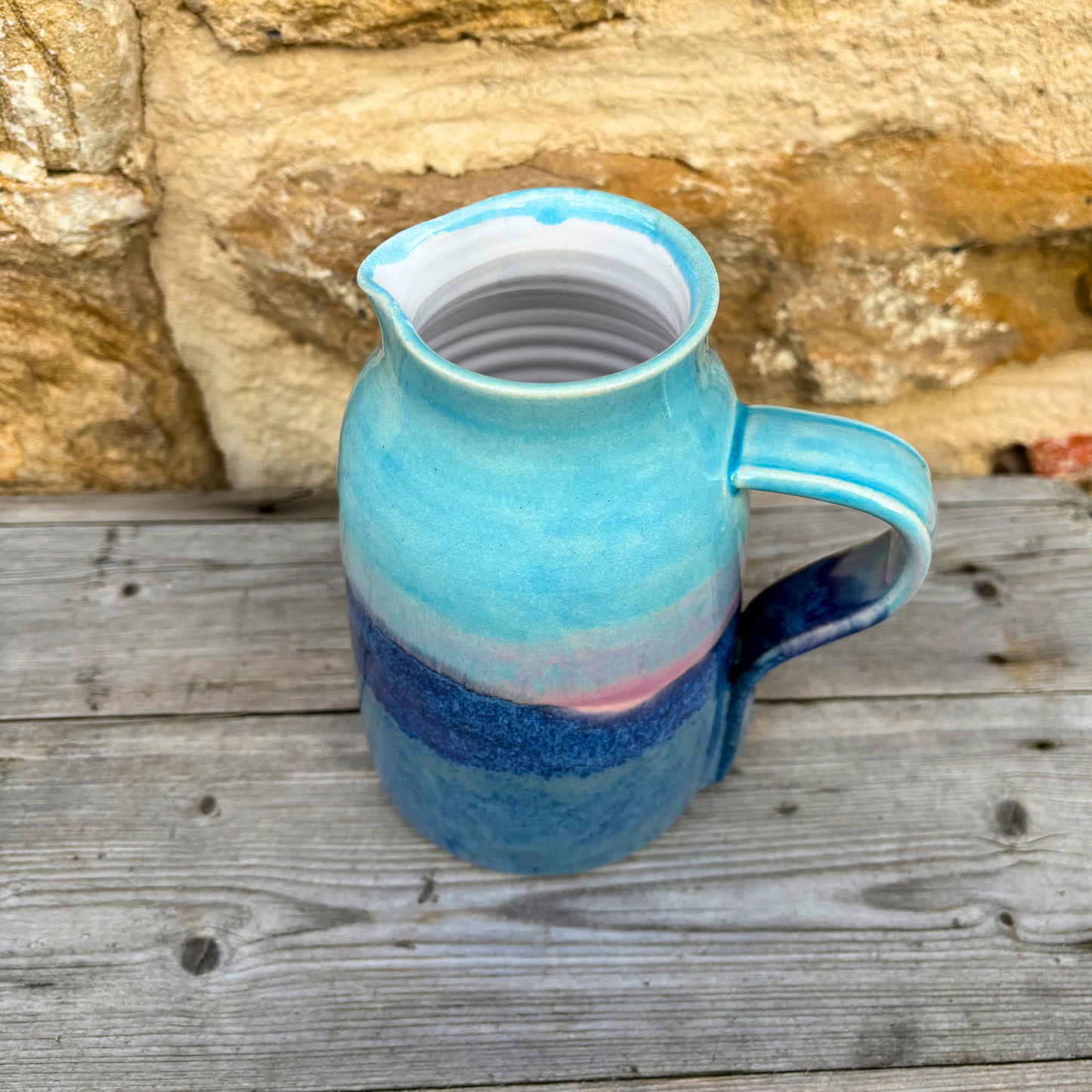 Blue ceramic pitcher with a white interior on a wooden surface with a stone wall background