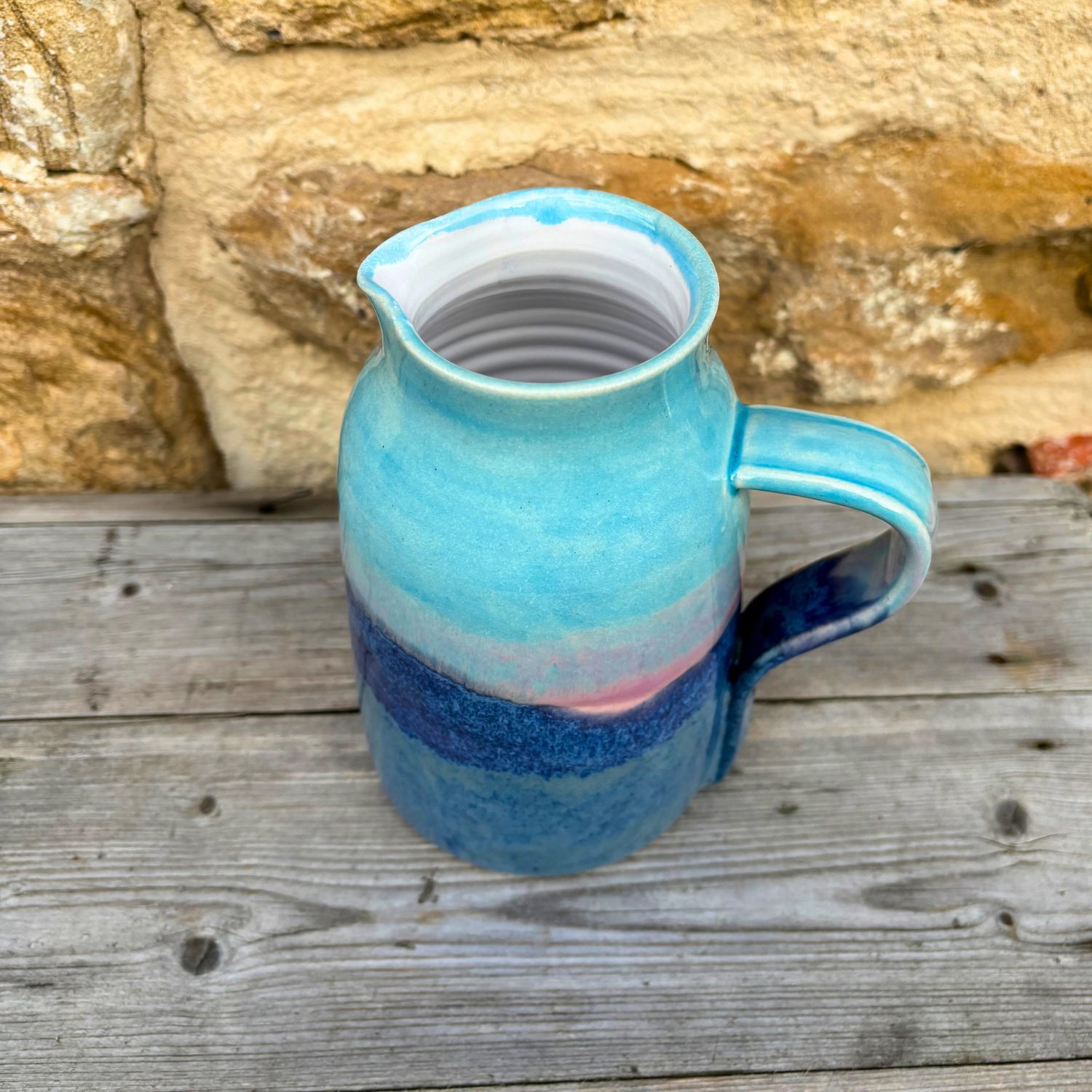 Blue ceramic pitcher with a white interior on a wooden surface with a stone wall background