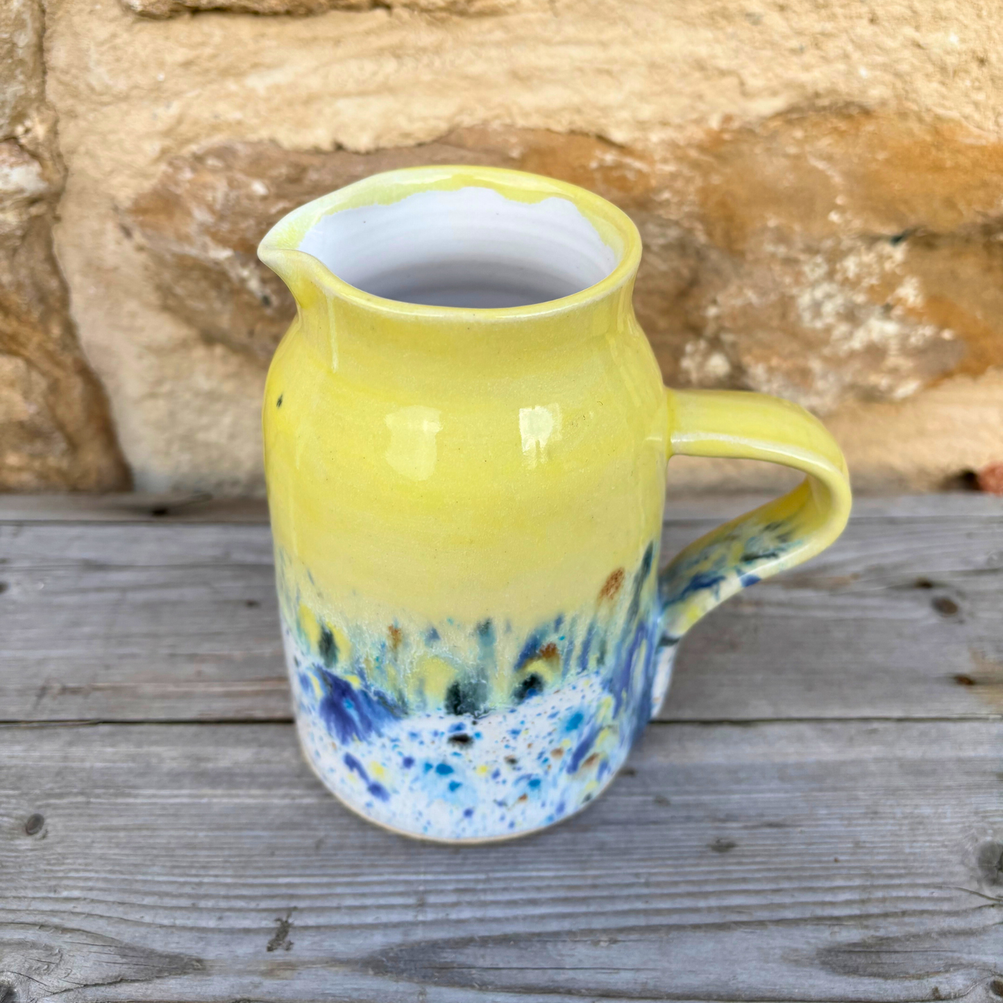 Yellow ceramic pitcher with blue and white speckled base on a wooden surface with stone background