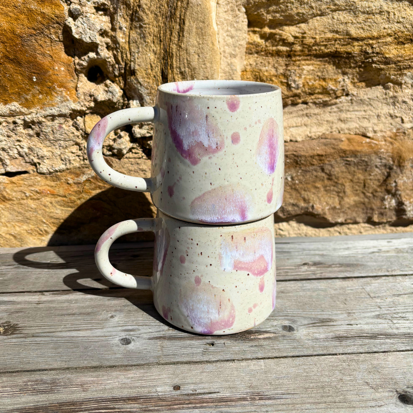 Stack of two ceramic mugs with pink speckles on a wooden surface with a stone wall background