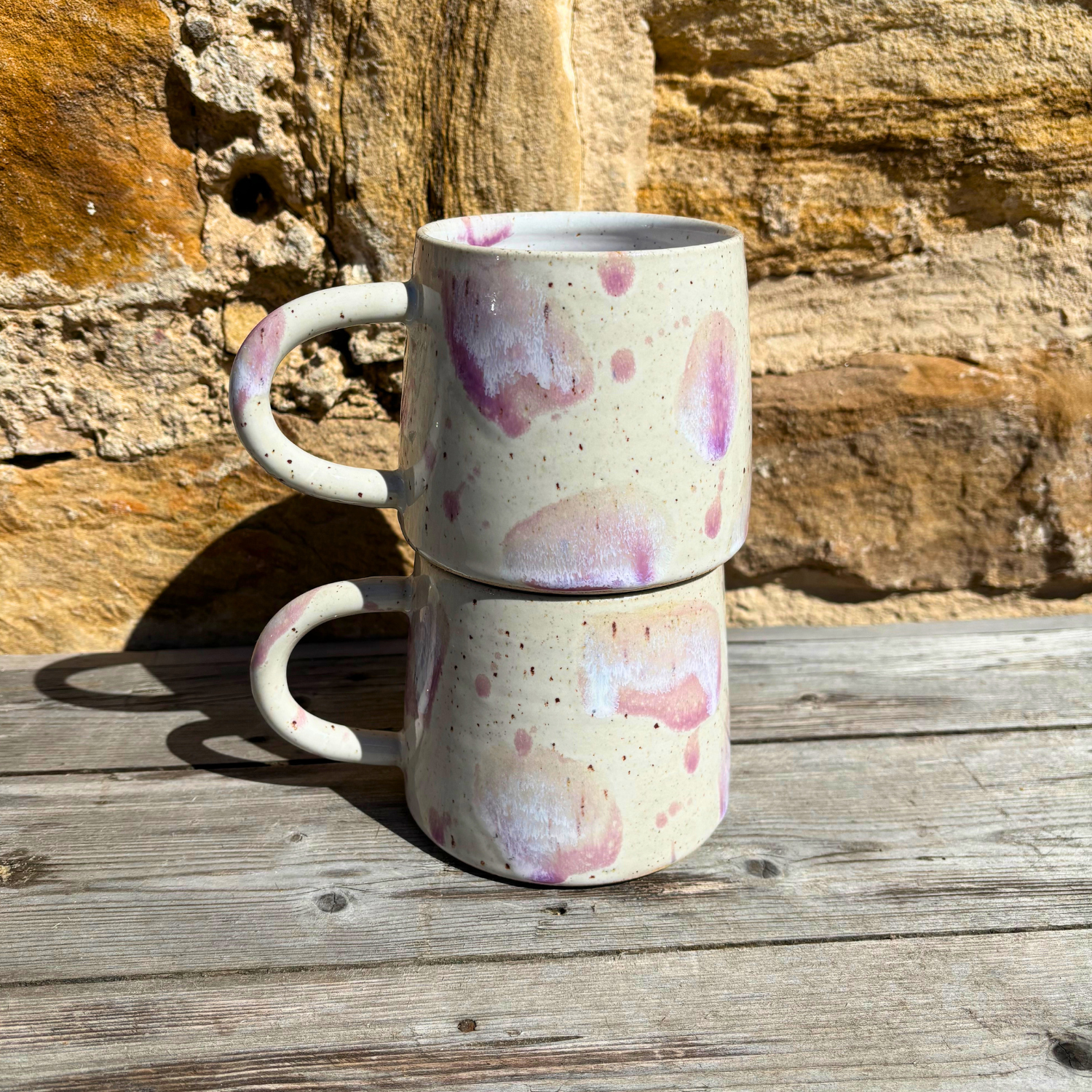 Stack of two ceramic mugs with pink speckles on a wooden surface with a stone wall background