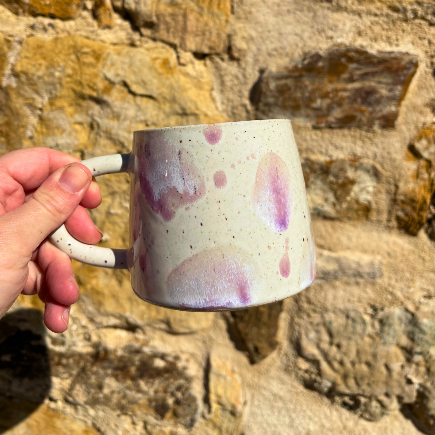 Hand holding a ceramic mug with purple speckles against a stone wall background