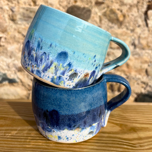 Two ceramic mugs with blue and multicolor glaze stacked on a wooden surface.