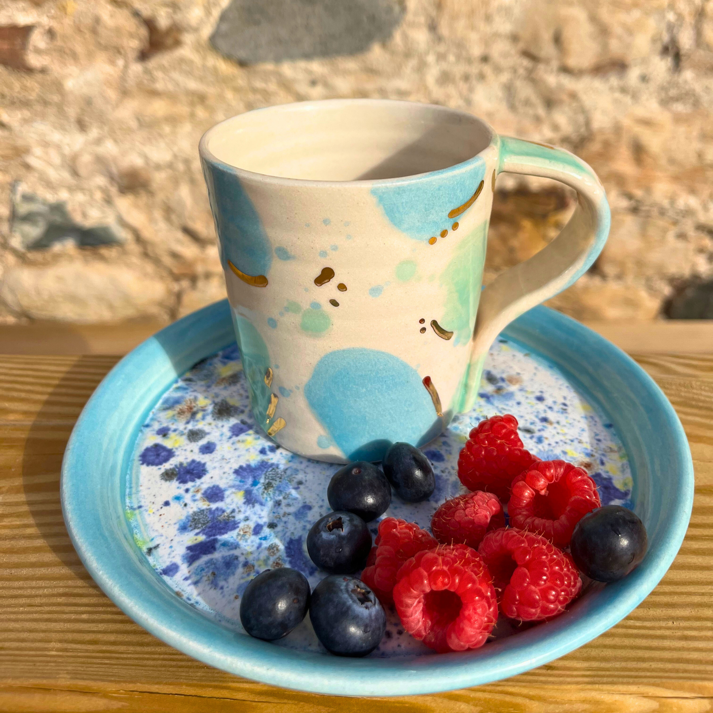 Ceramic mug with blue and beige design on a blue plate with berries against a stone wall background