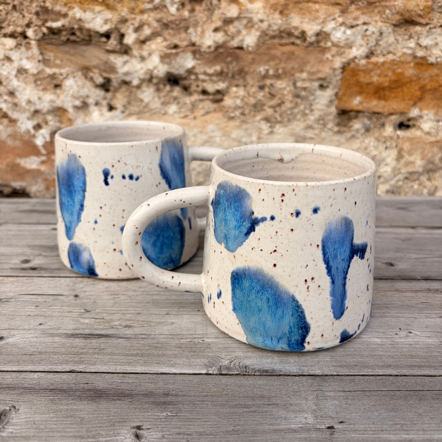 Two ceramic mugs with blue abstract designs on a wooden surface with a stone wall background.