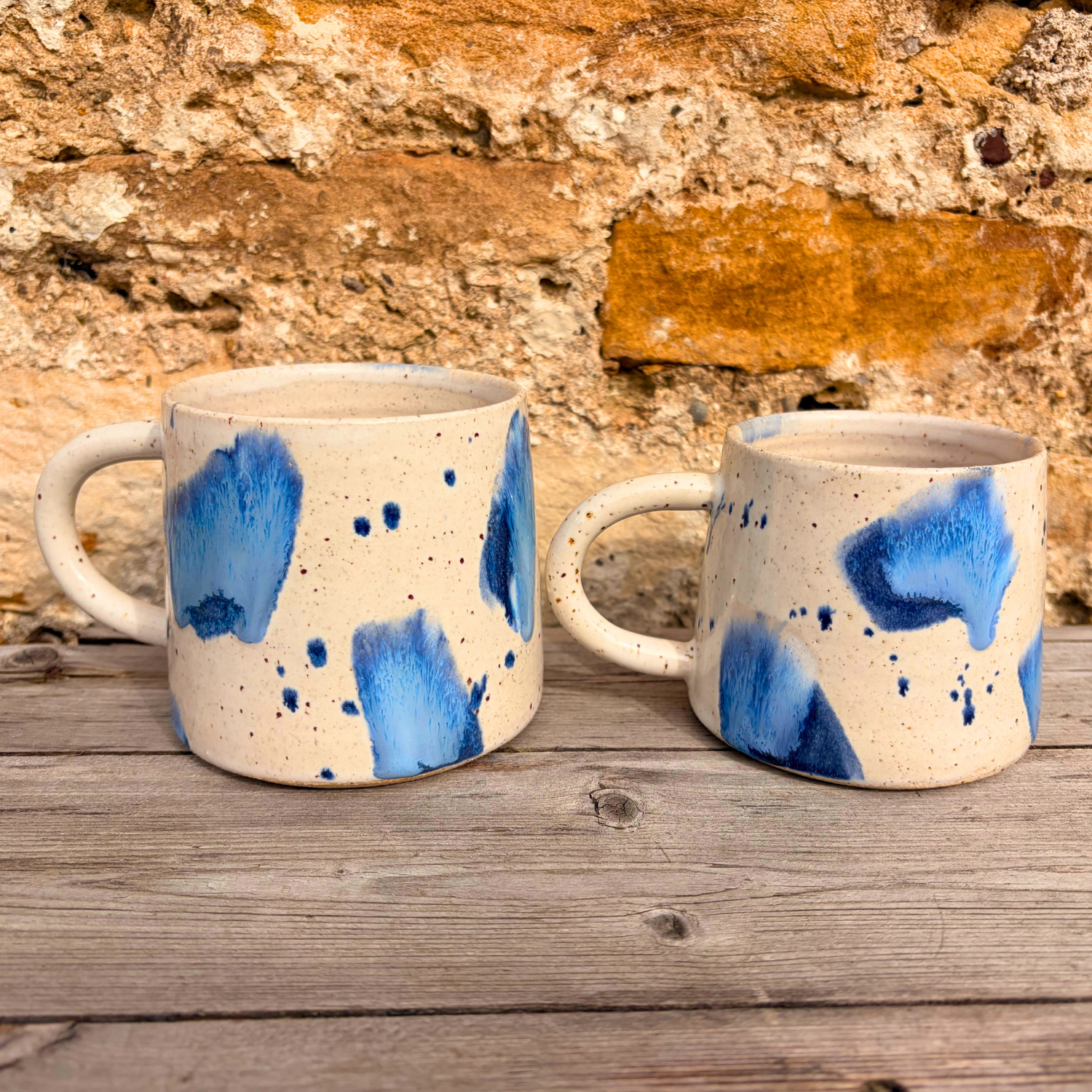 Two ceramic mugs with blue abstract patterns on a wooden surface with a stone wall background.