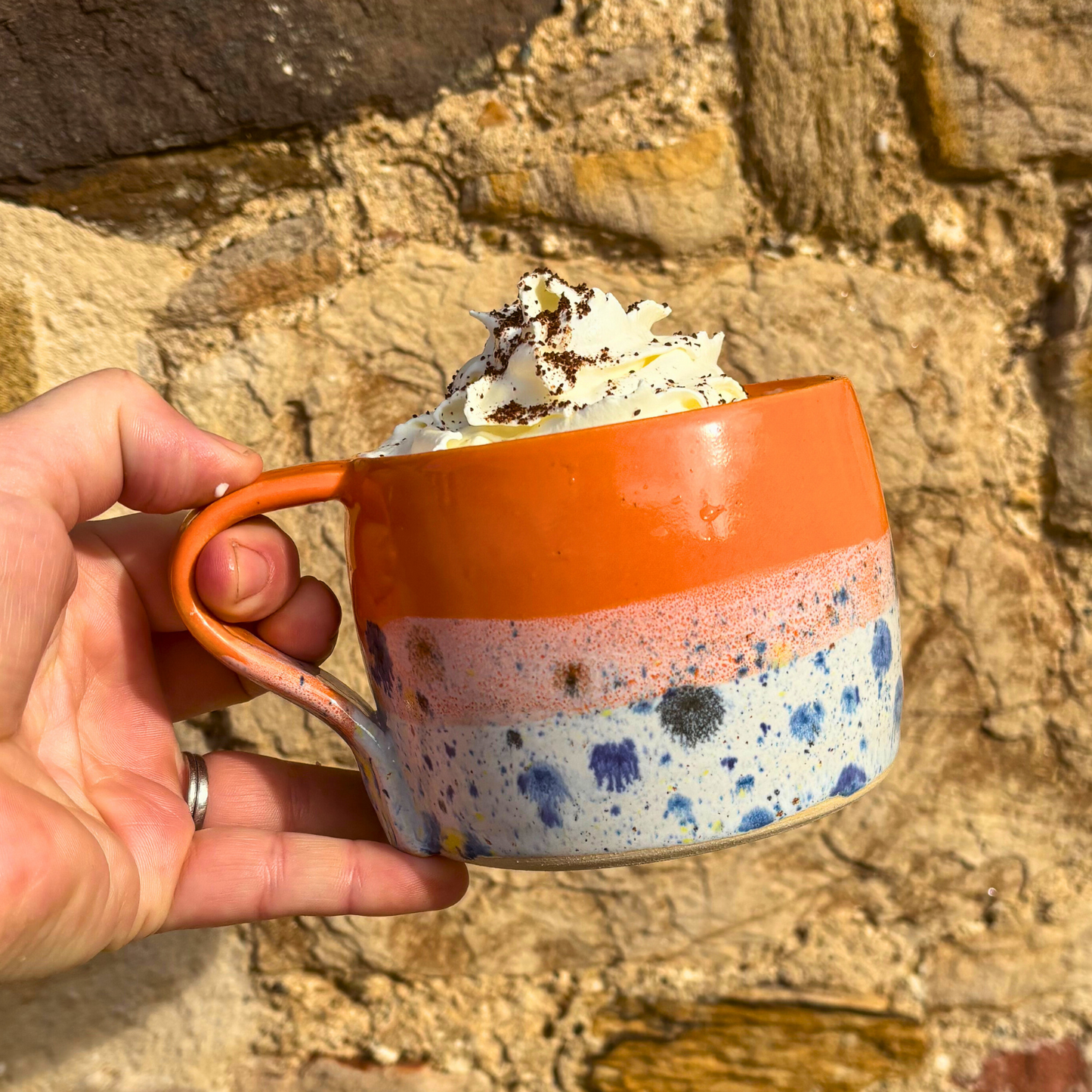 Hand holding a colorful mug with whipped cream against a stone background