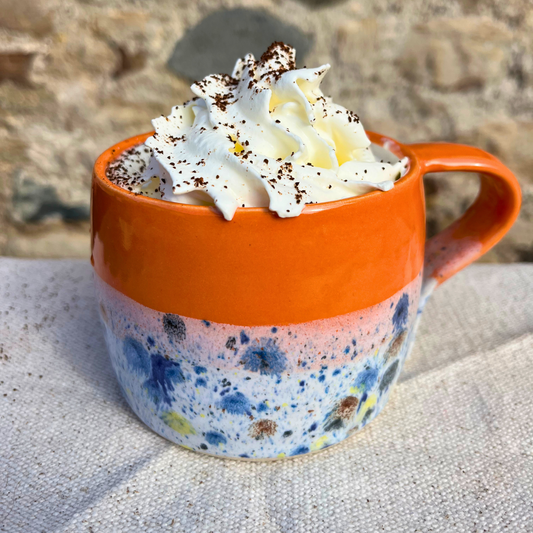 Orange mug with floral pattern filled with hot chocolate and whipped cream, set against a stone wall background.
