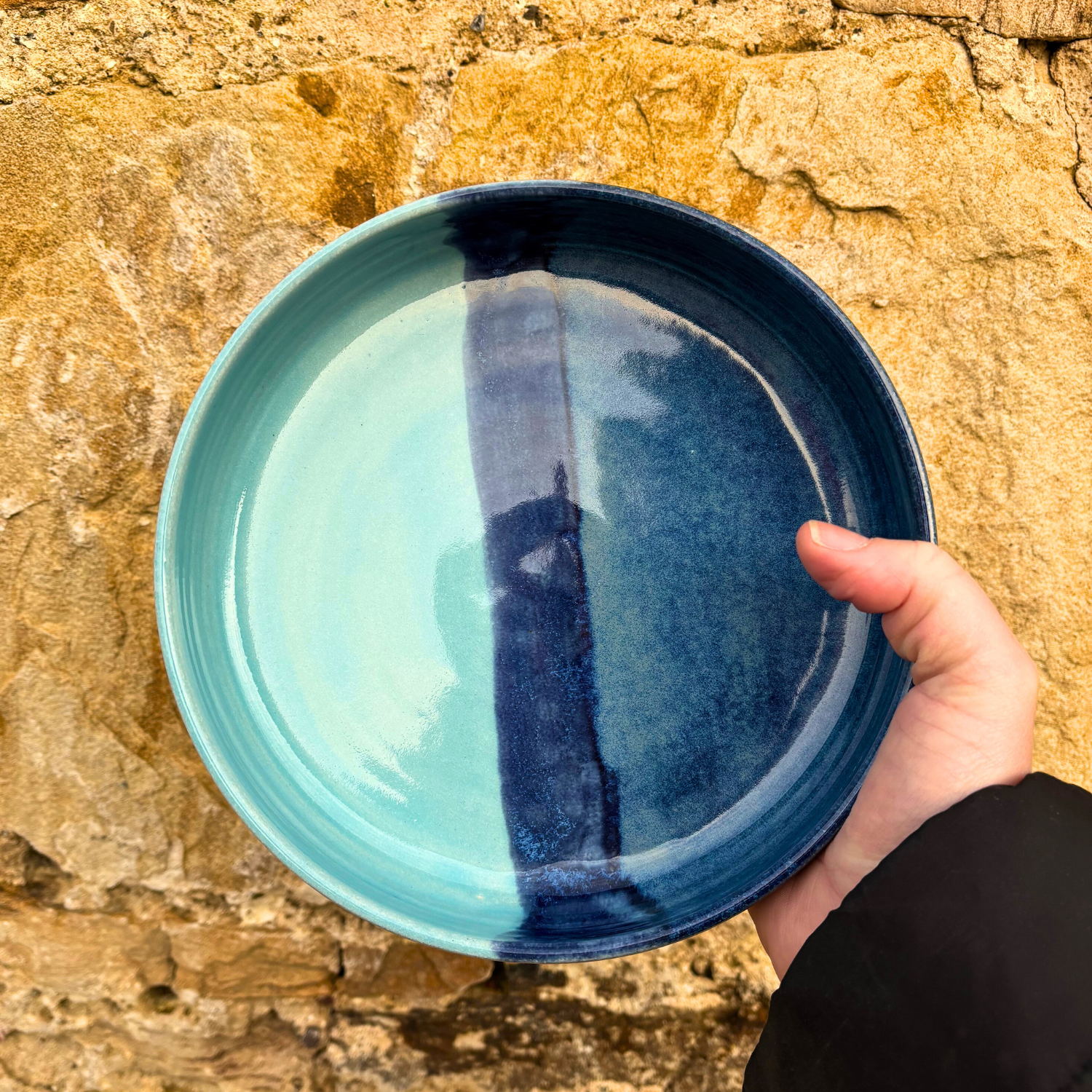 Hand holding a blue ceramic plate against a stone wall.