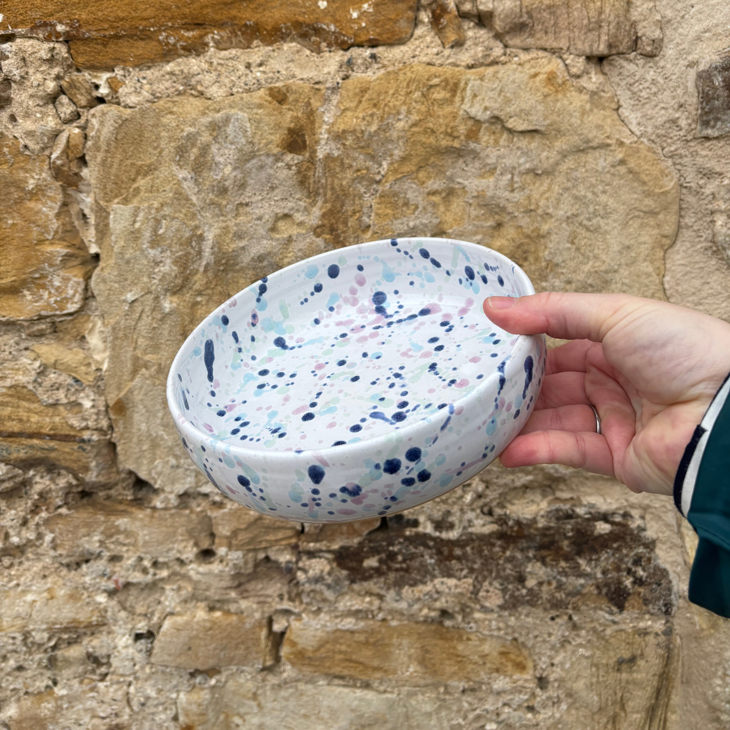 Hand holding a ceramic bowl with blue and pink speckles against a stone wall background