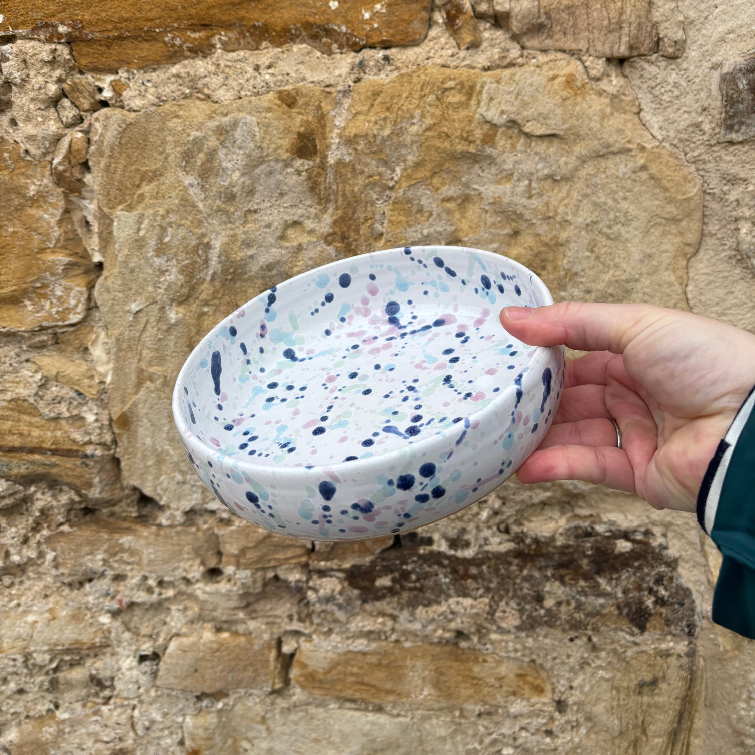 Hand holding a ceramic bowl with blue and pink speckles against a stone wall background