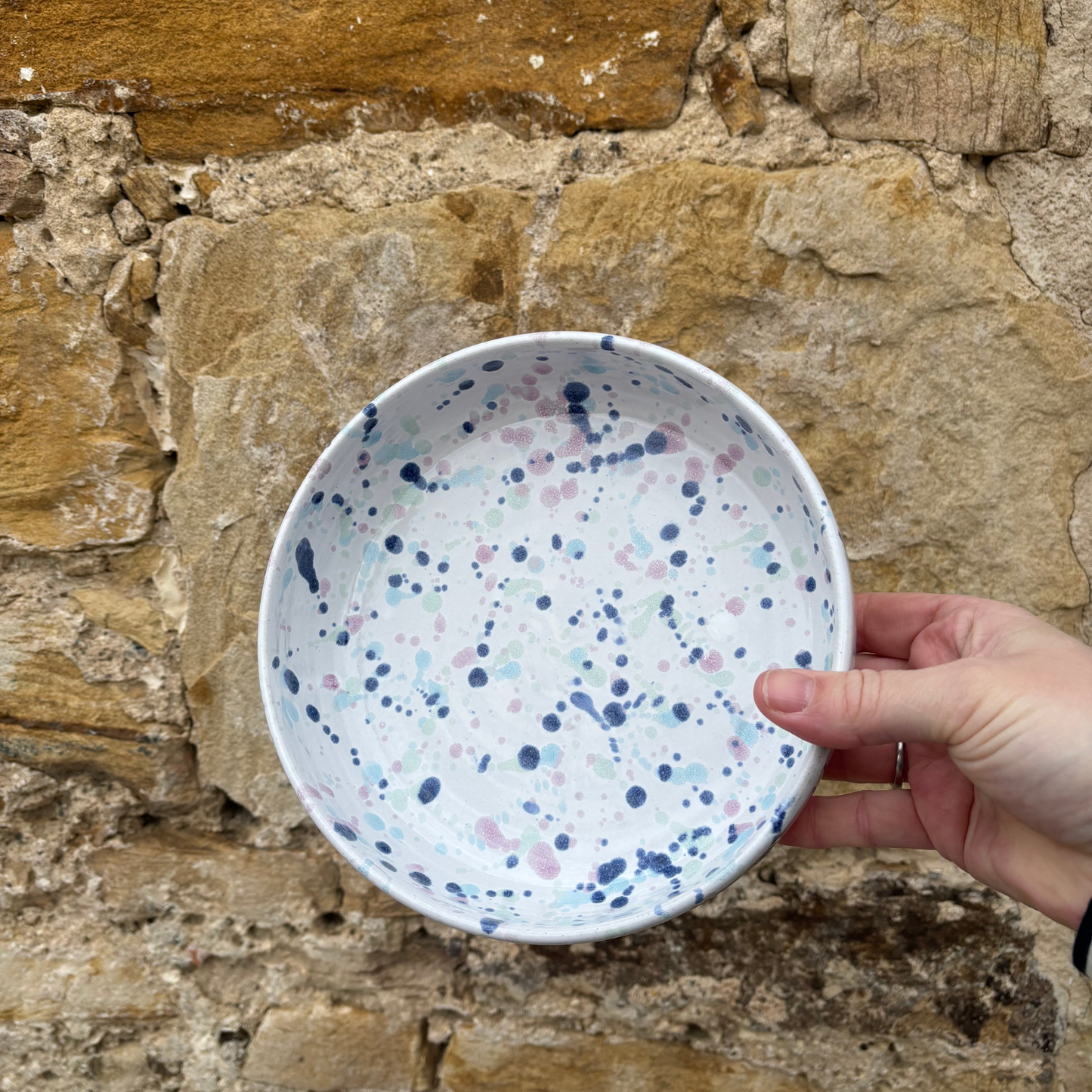 Hand holding a ceramic bowl with blue and pink speckles against a stone wall background