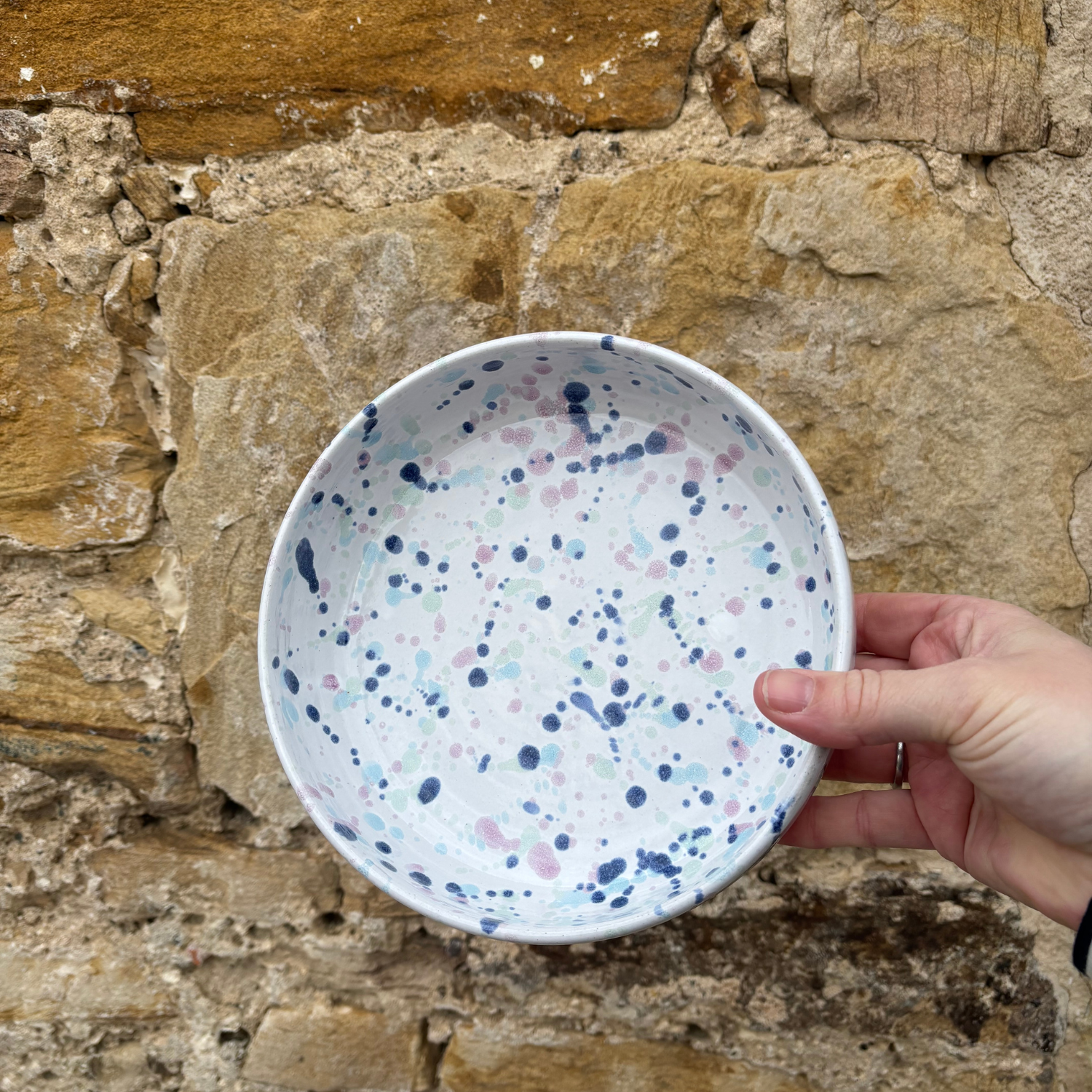Hand holding a ceramic bowl with blue and pink speckles against a stone wall background