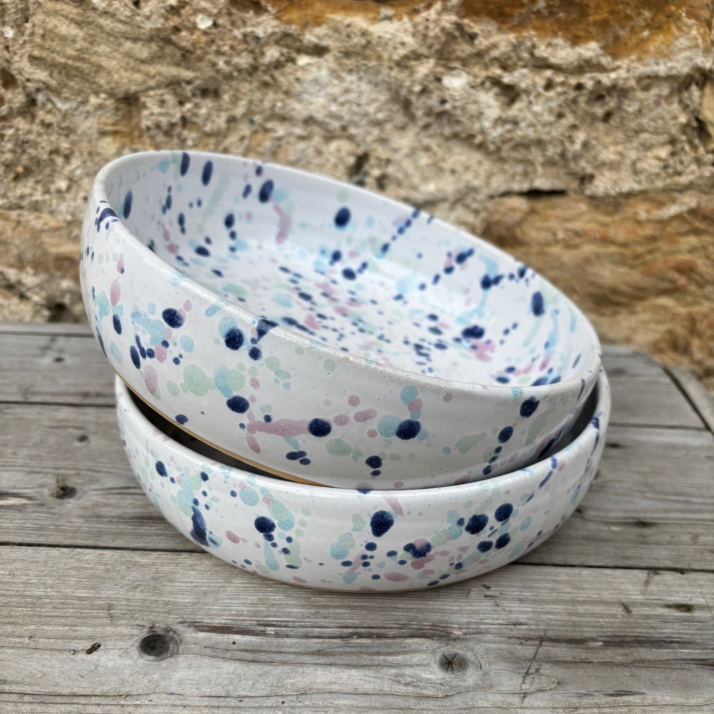 Two ceramic bowls with a speckled pattern stacked on a wooden surface with a stone wall background.