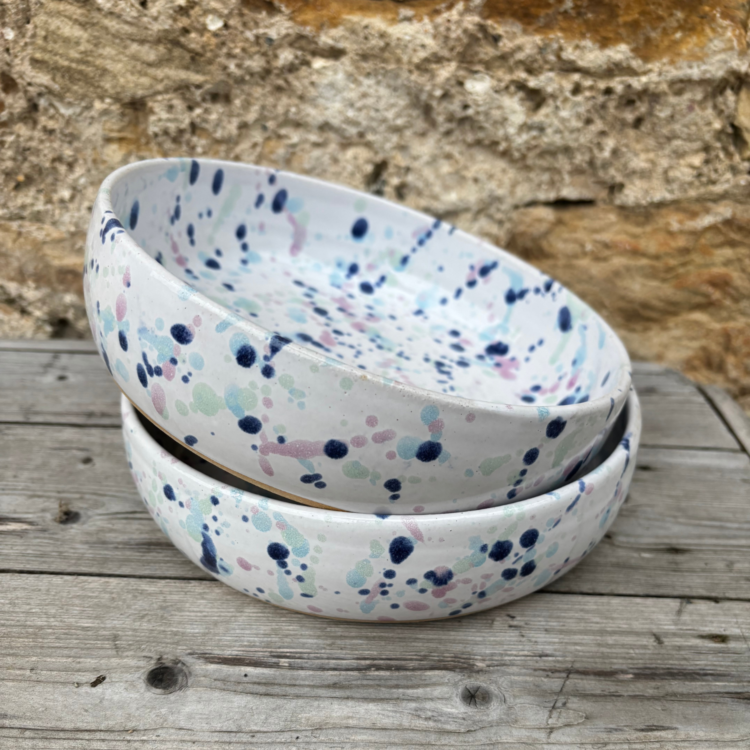 Two ceramic bowls with a speckled pattern stacked on a wooden surface with a stone wall background.