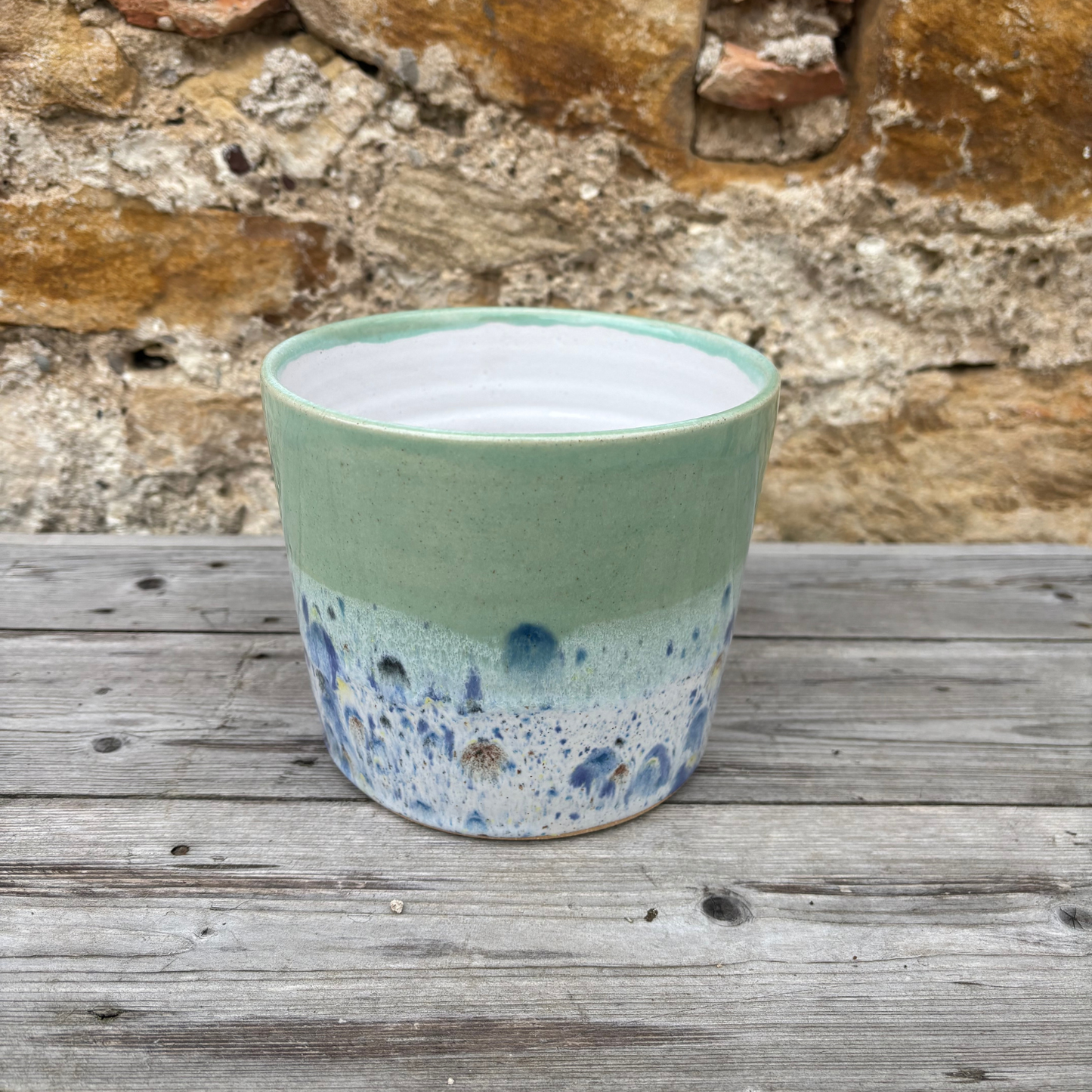 Green ceramic pot with blue speckles on a wooden surface with a stone wall background